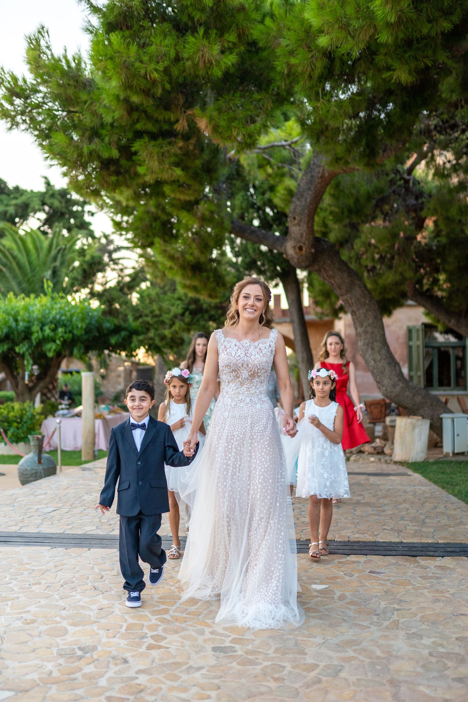 Wedding party walks along stone pathway surrounded by green trees.