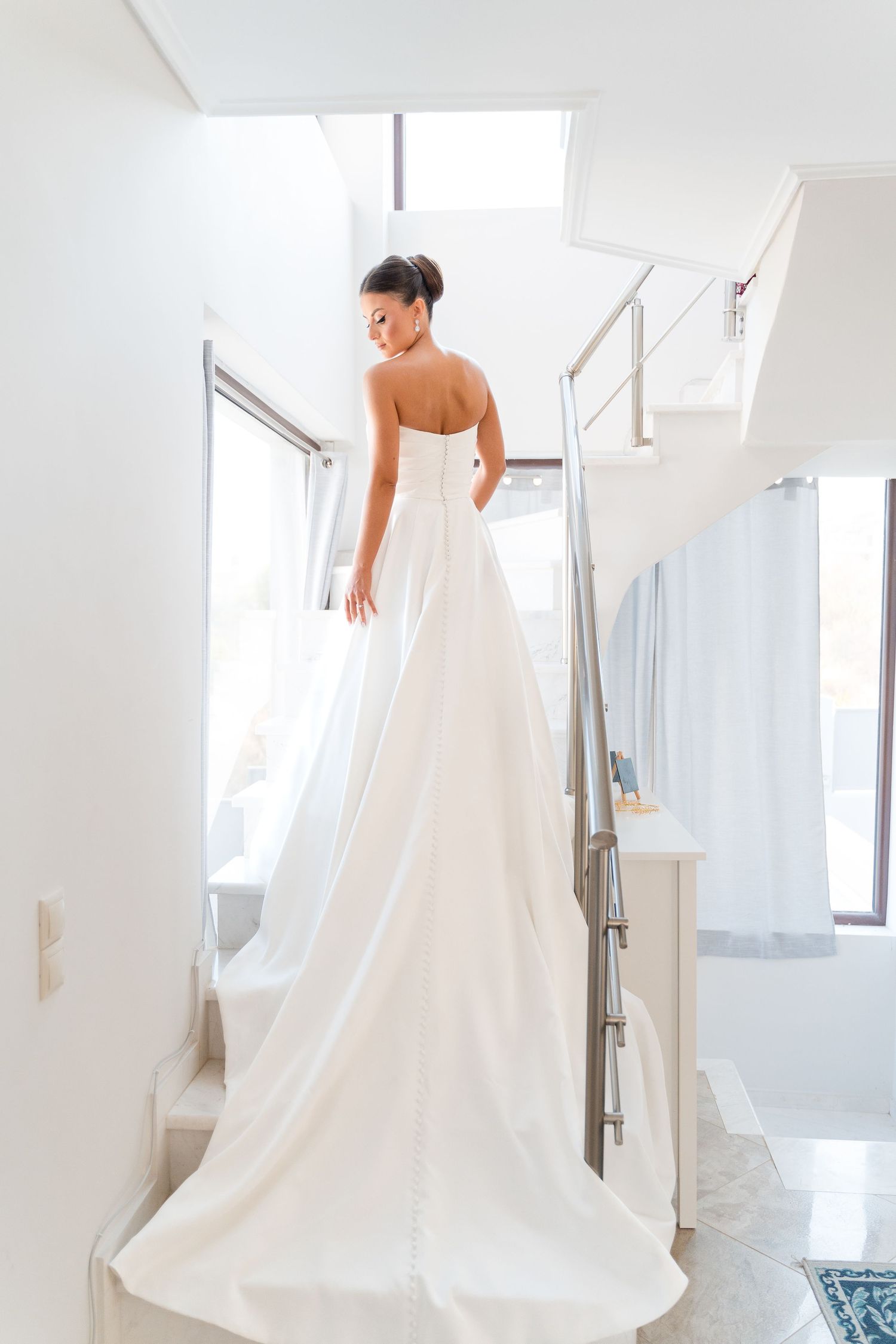 A bride in a flowing white strapless wedding gown stands on a bright marble staircase with modern railings.