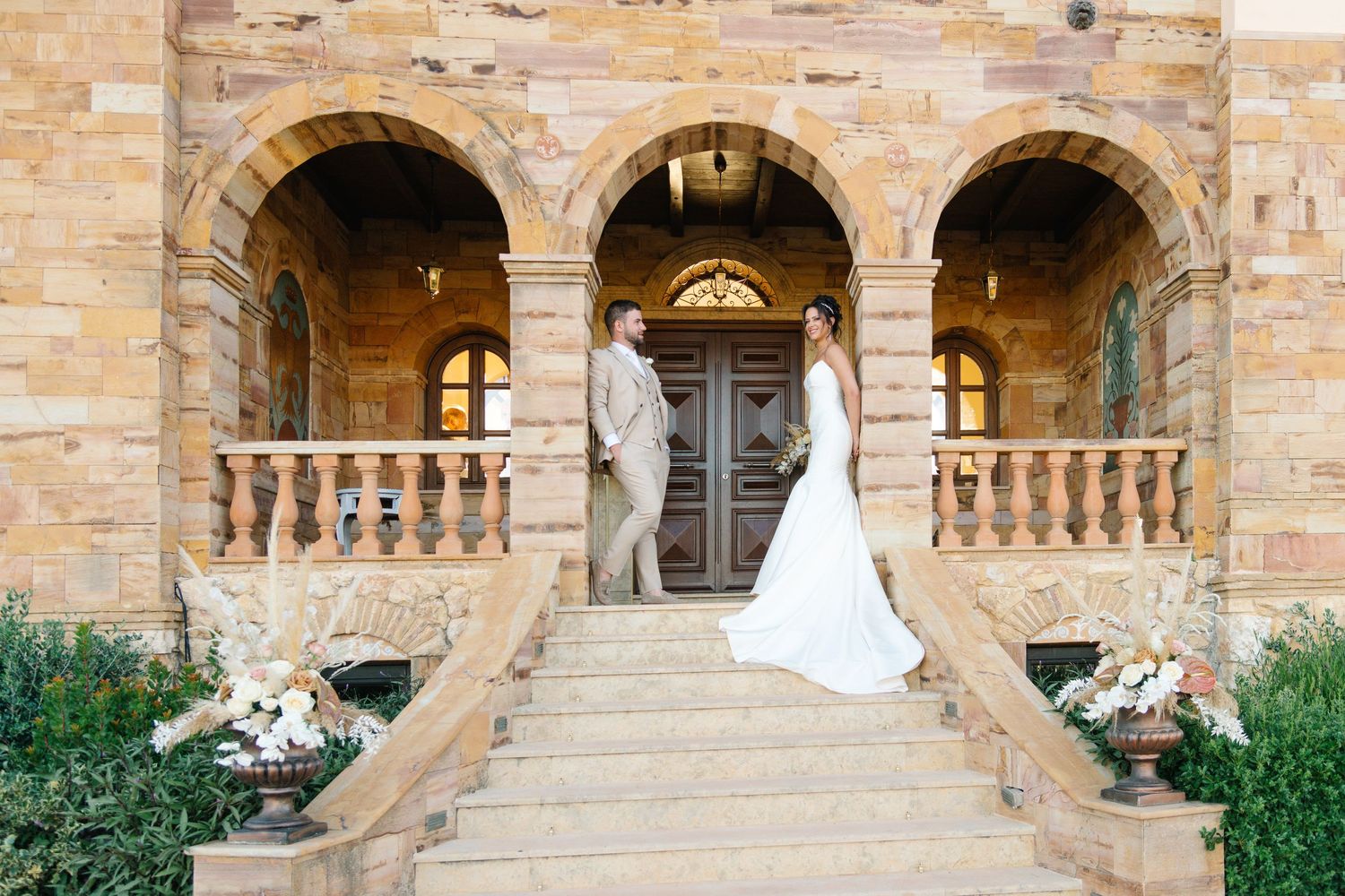 Bride and groom stand on stone staircase of elegant villa with arched architecture on their wedding day.