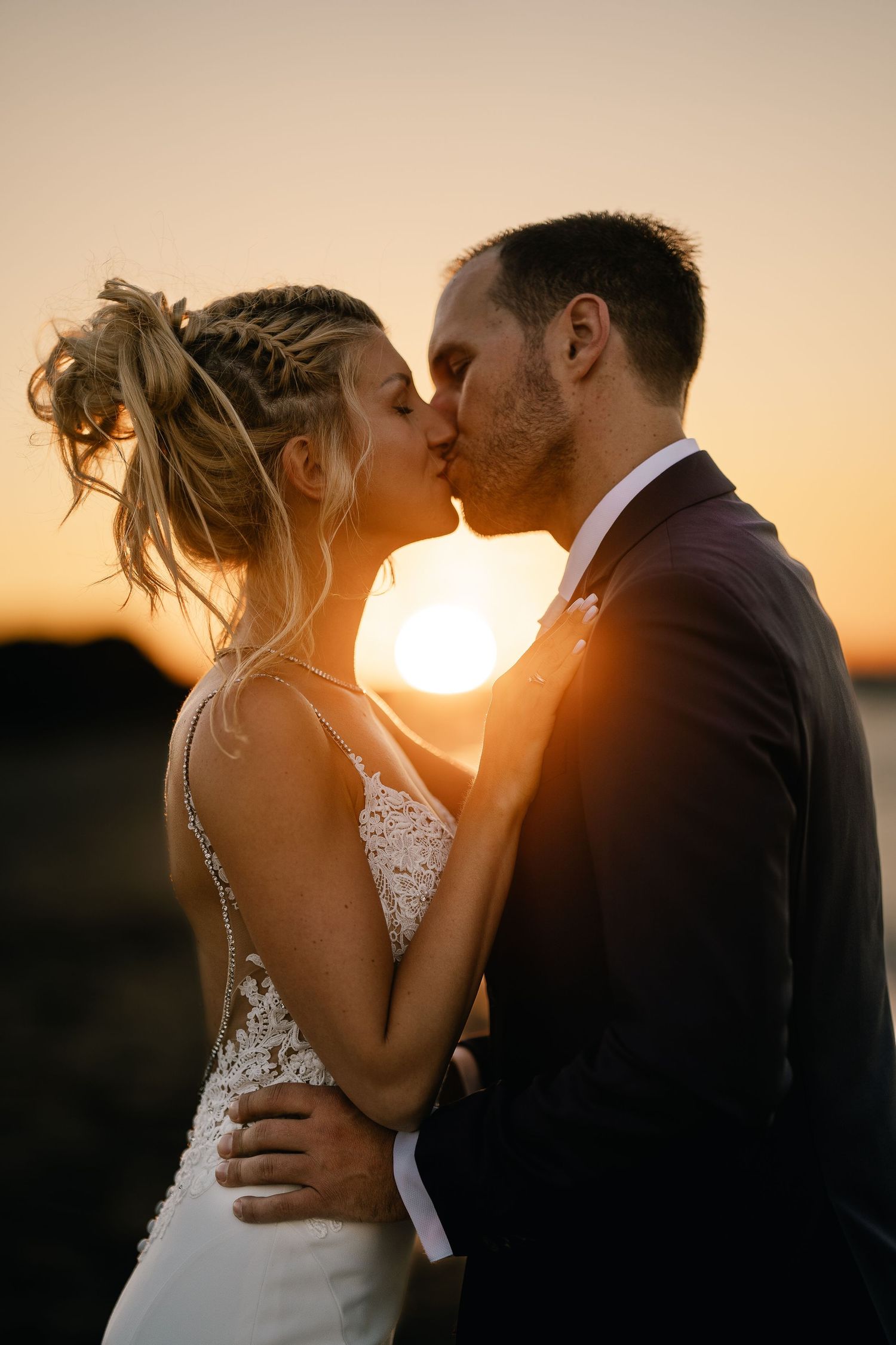 A wedding couple shares an intimate kiss at sunset creating a romantic silhouette against the golden sky.