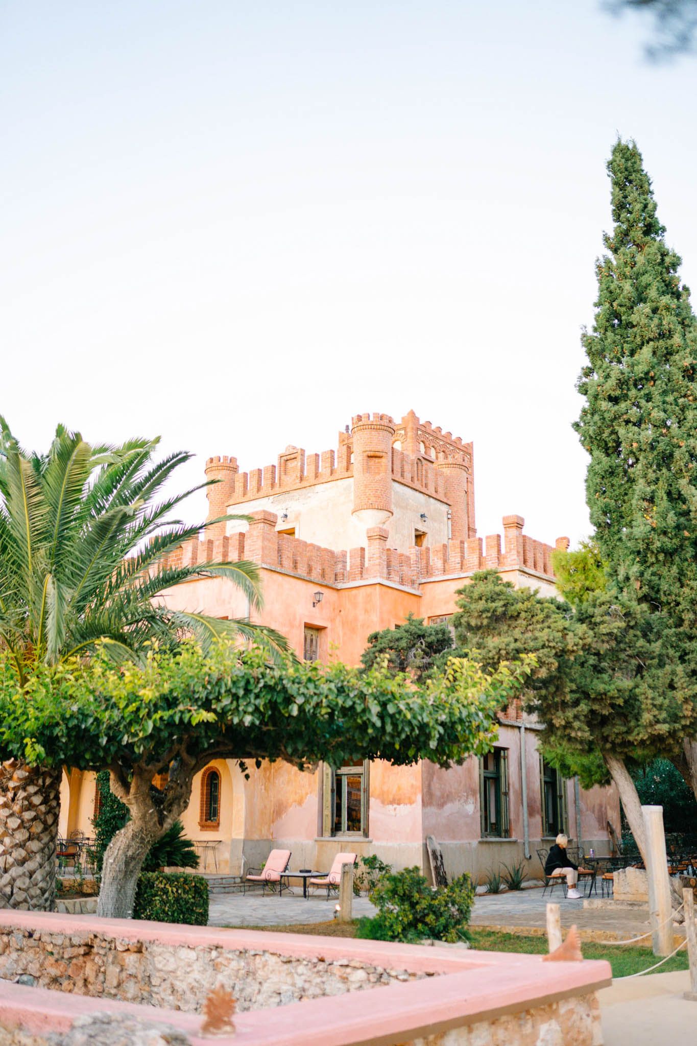 A Mediterranean-style villa with terracotta walls and castle-like architecture surrounded by lush gardens and palm trees.