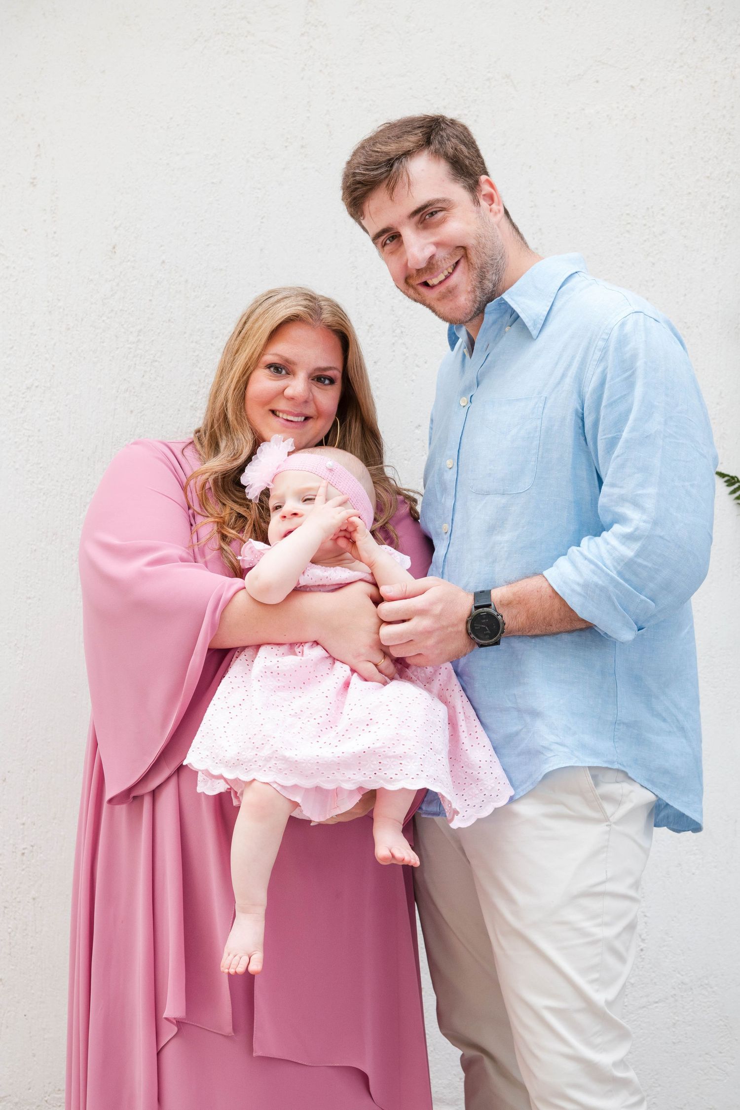 A family poses together for portrait photos in pink and blue outfits with a sleeping baby in a white background setting.