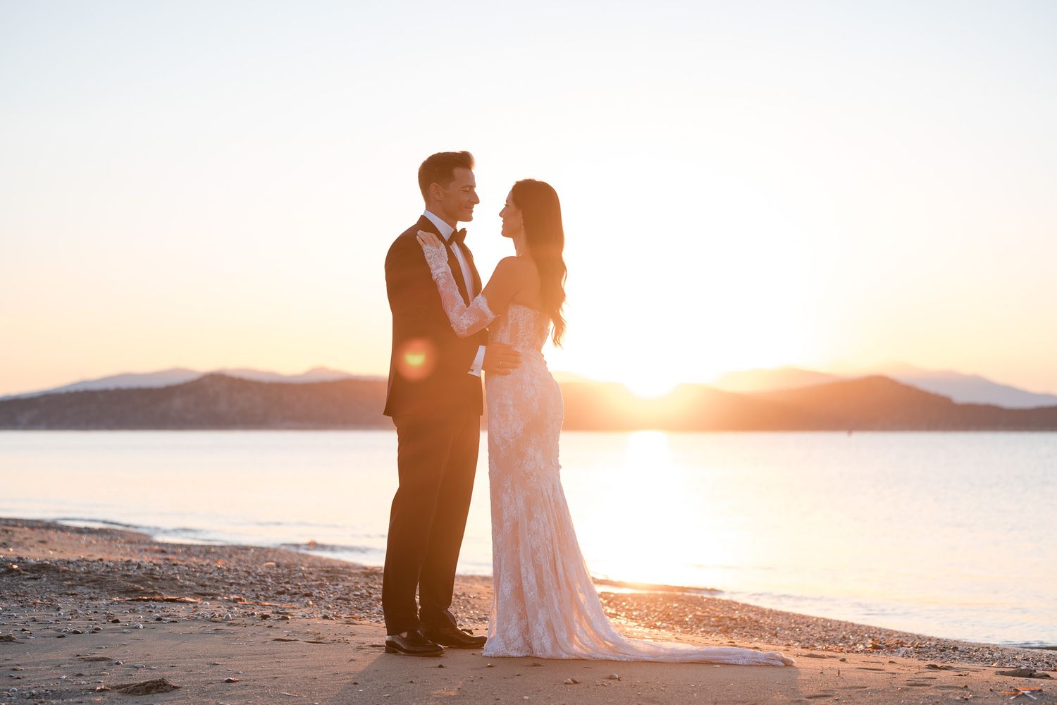 Bride and groom share a romantic sunset moment on a beach, with golden light and mountains reflecting on calm water.