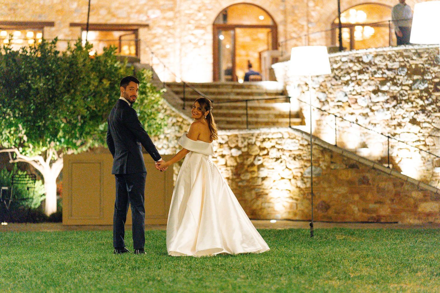 Bride and groom hold hands at night outside a stunning stone villa, glowing with warm lights.