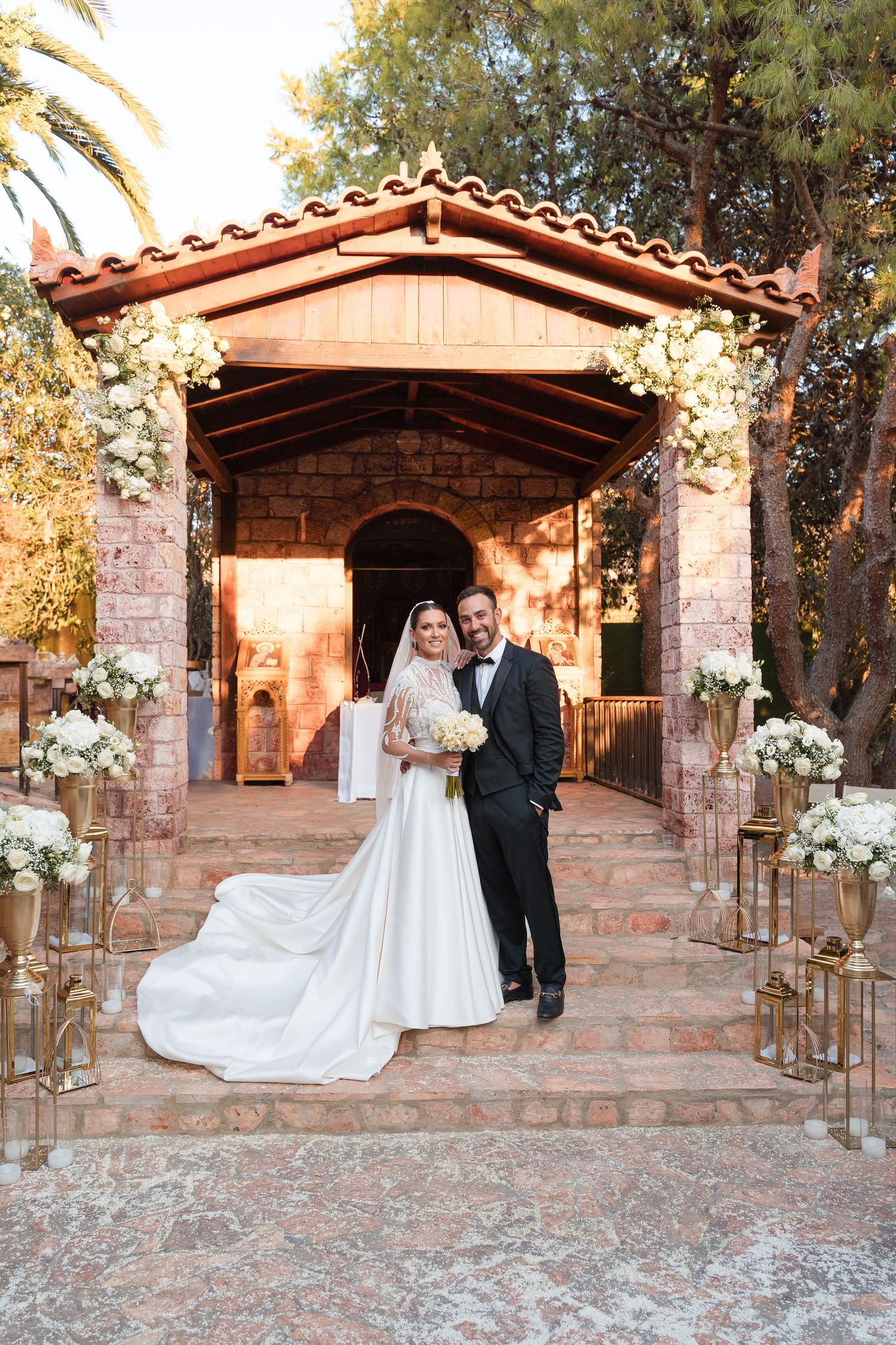 Elegant outdoor wedding ceremony at a rustic stone chapel with terracotta roof and white floral decorations along the aisle.