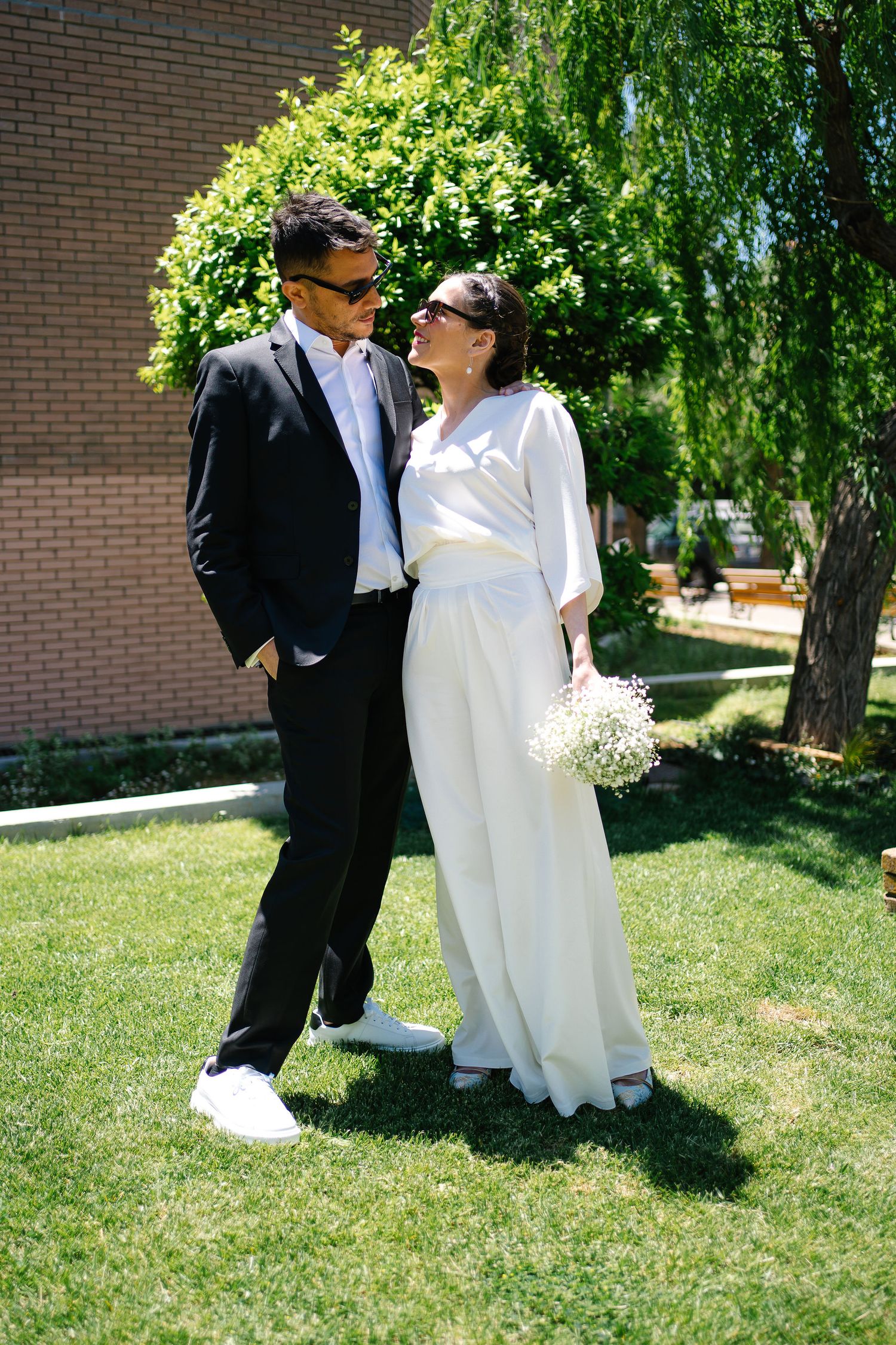 A couple shares joyful moments outdoors on their wedding day against a backdrop of lush greenery and brick walls.
