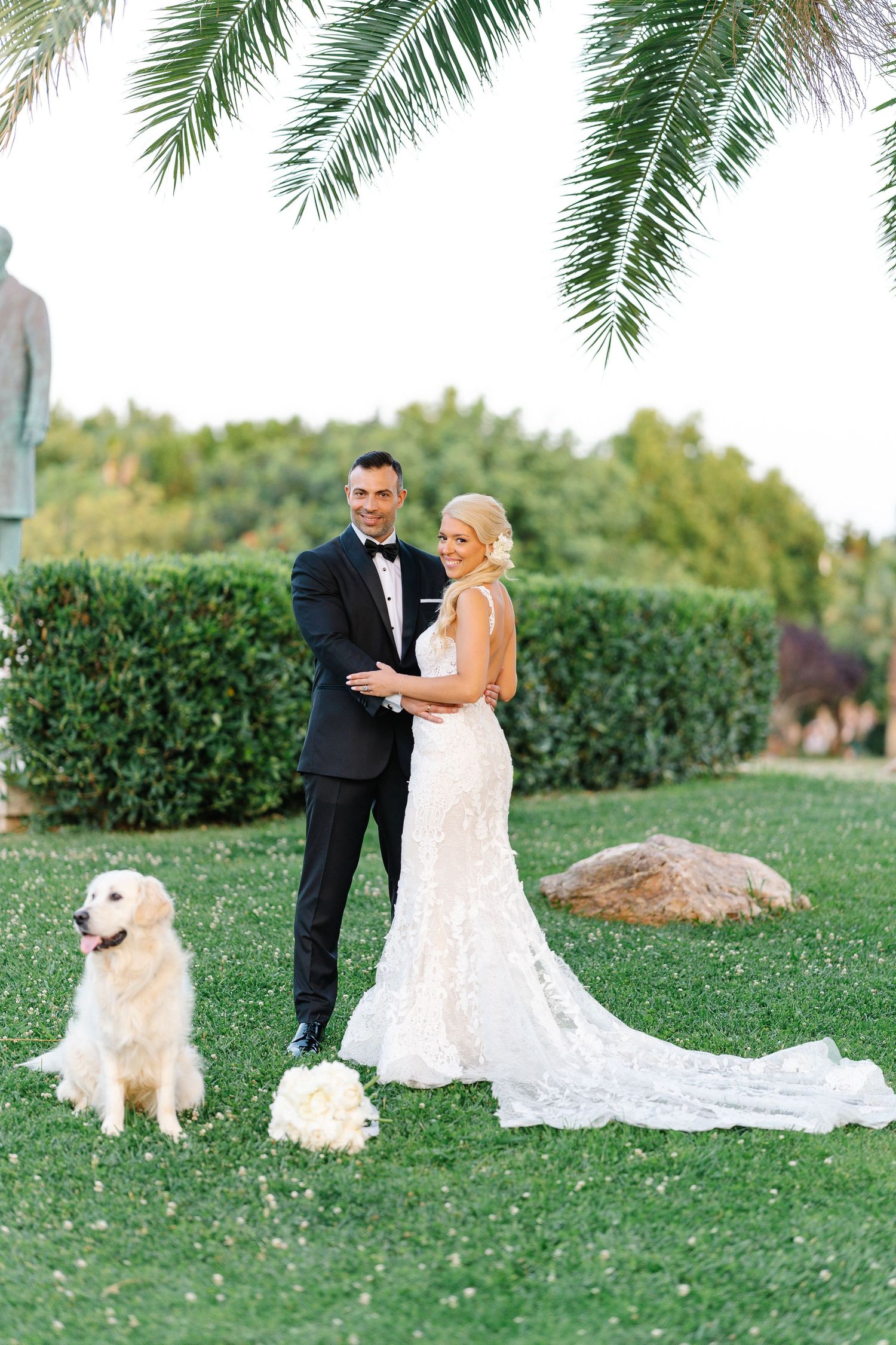 Wedding couple poses with golden retriever dog on manicured lawn under palm trees.