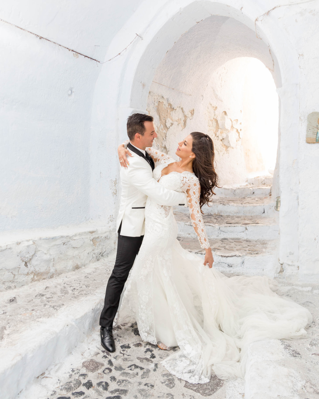 A couple dancing and twirling in elegant wedding attire against a rustic white stone backdrop with arched passageways.
