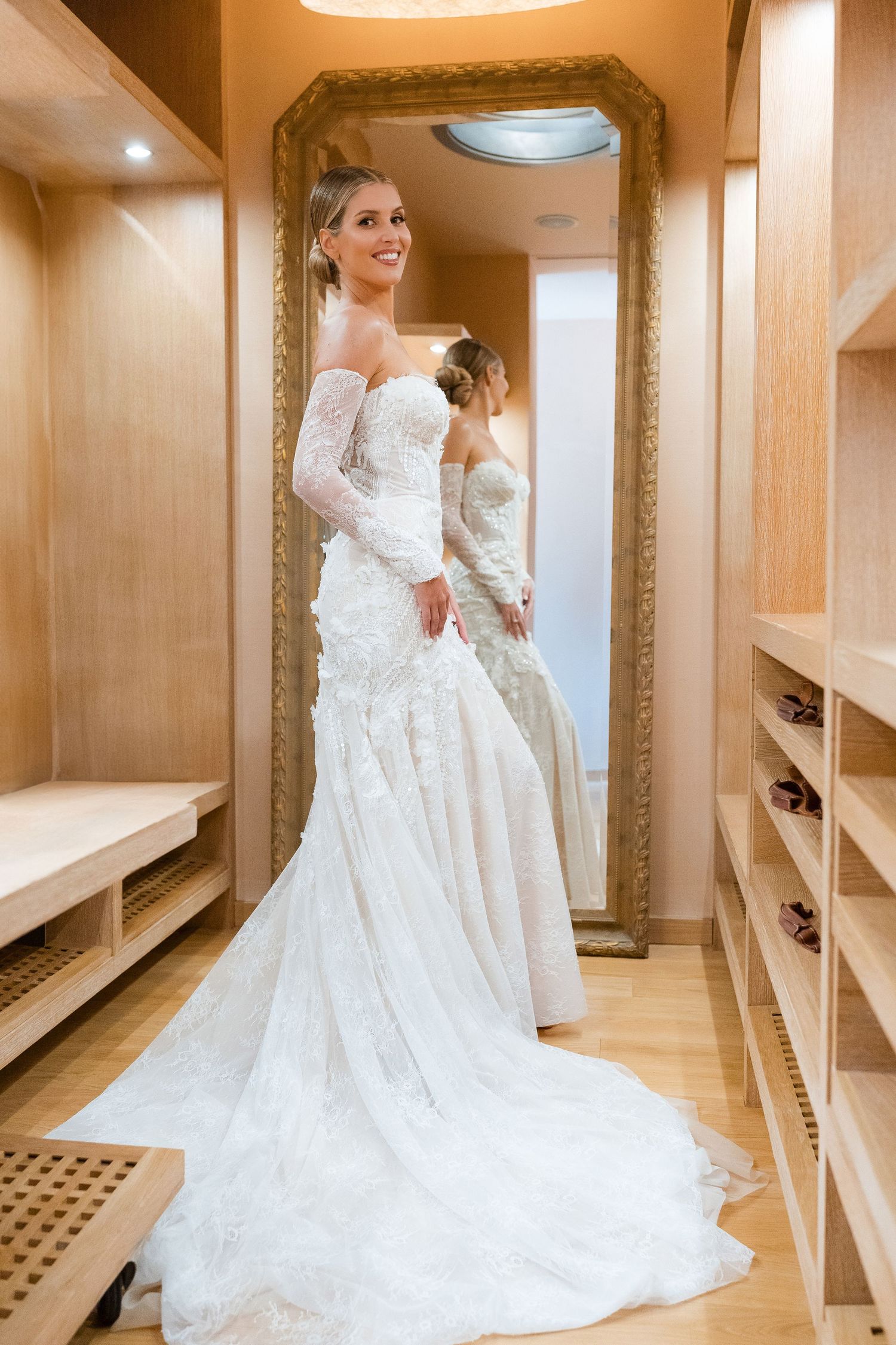 A white lace wedding dress with long sleeves and train displayed in an elegant dressing room with mirrored walls.