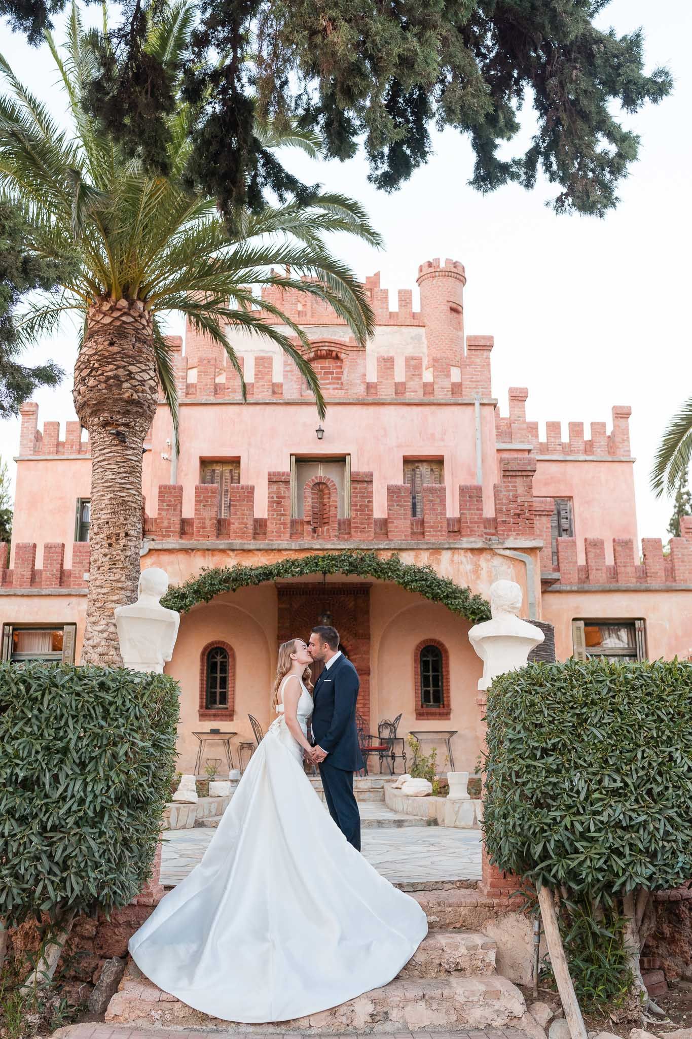 Elegant wedding portraits in front of pink castle-like building with palm trees.