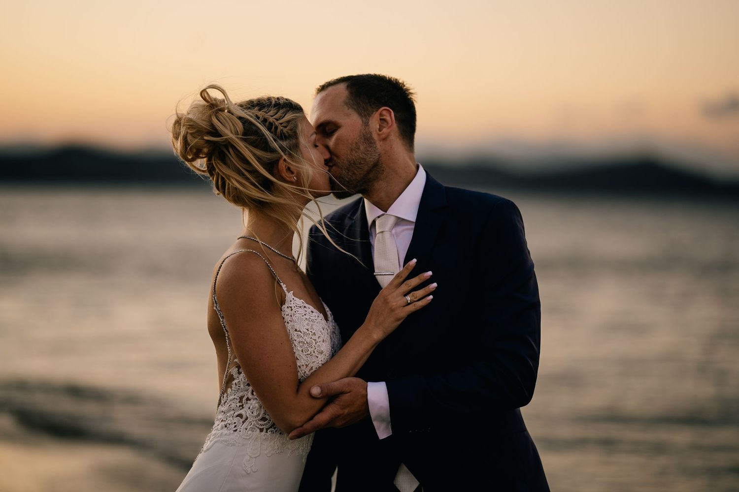 Bride and groom share a romantic sunset kiss on the beach, with golden hues reflecting over calm ocean waters.