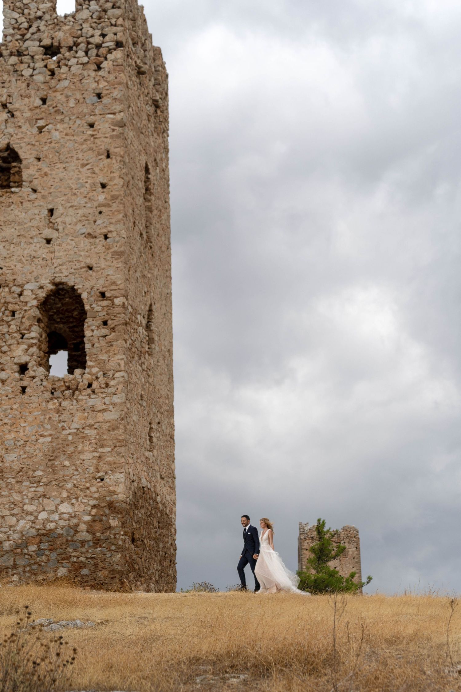 A couple stands near ancient stone ruins and tower against a cloudy sky in a romantic wedding photoshoot.