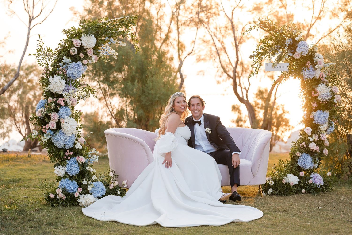 Elegant outdoor wedding setup with blue floral arch and white loveseat at sunset in a garden setting.