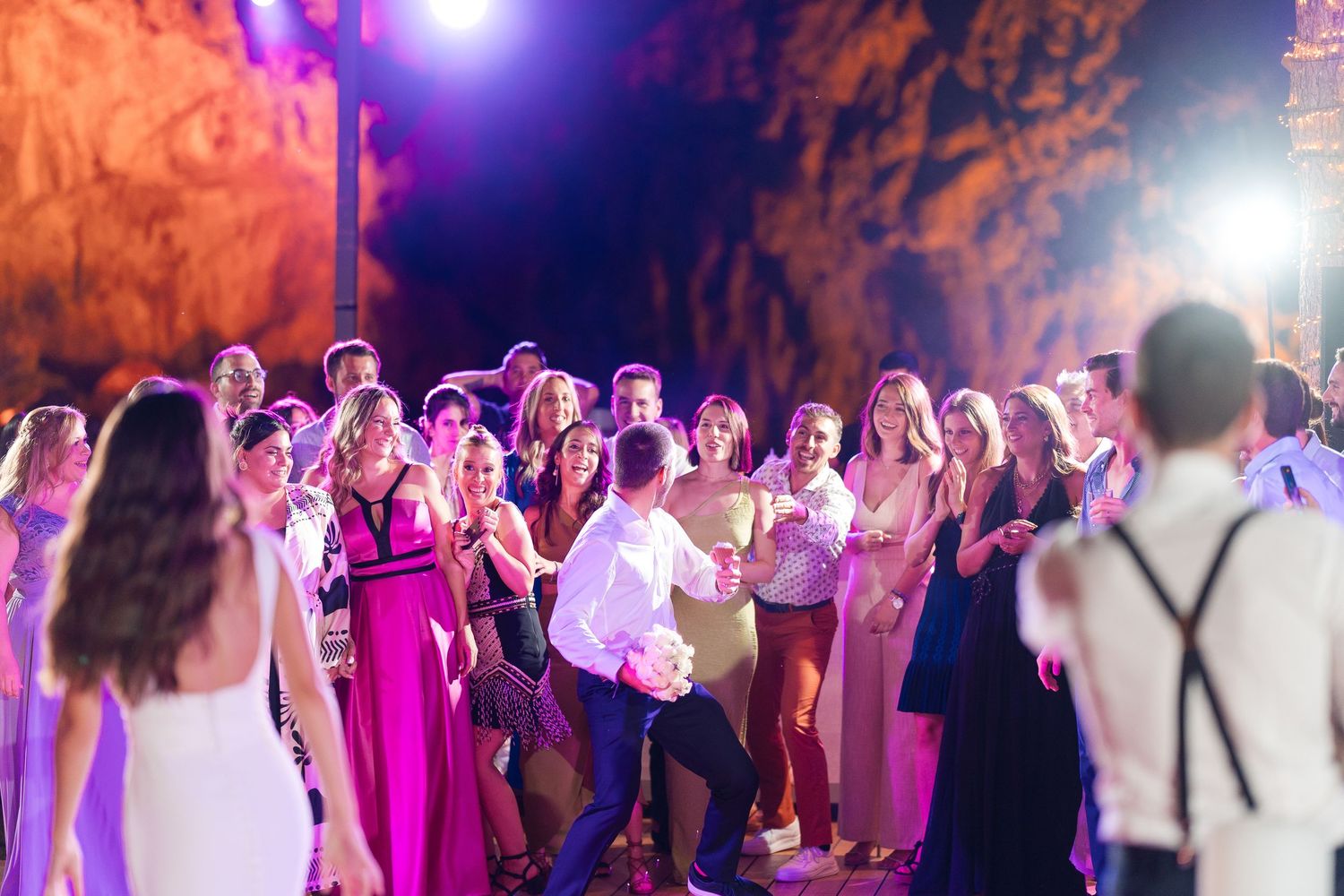 Wedding reception guests dance together on a crowded dance floor illuminated by purple and blue party lights.