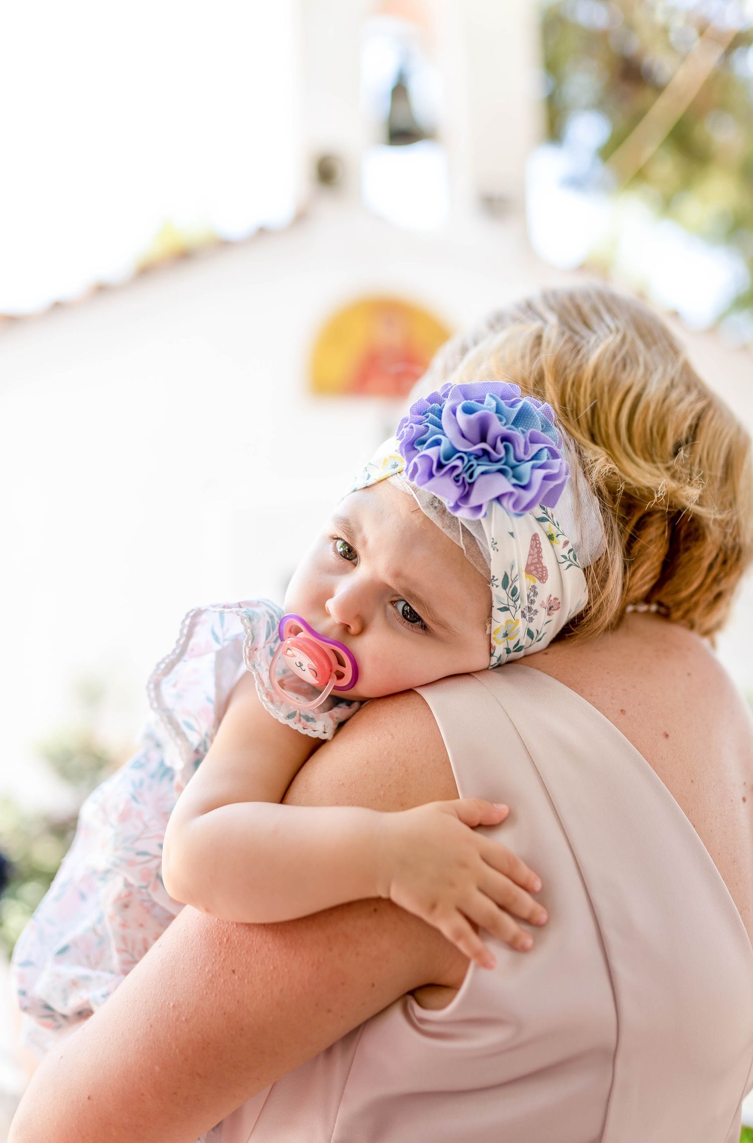 Sweet baby wearing floral headband rests head on shoulder while holding pacifier at outdoor event.