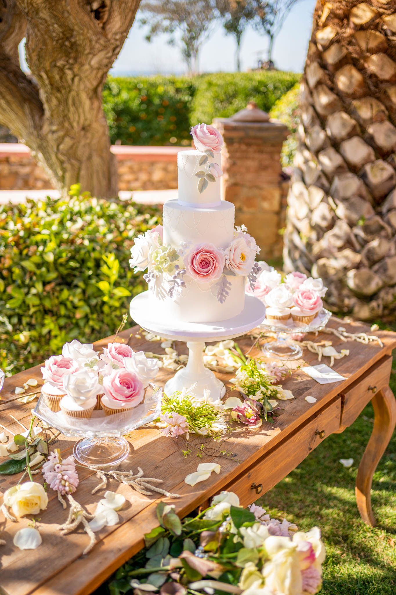 An elegant wedding dessert table decorated with white cakes, pink roses and rustic wooden elements in a garden setting.
