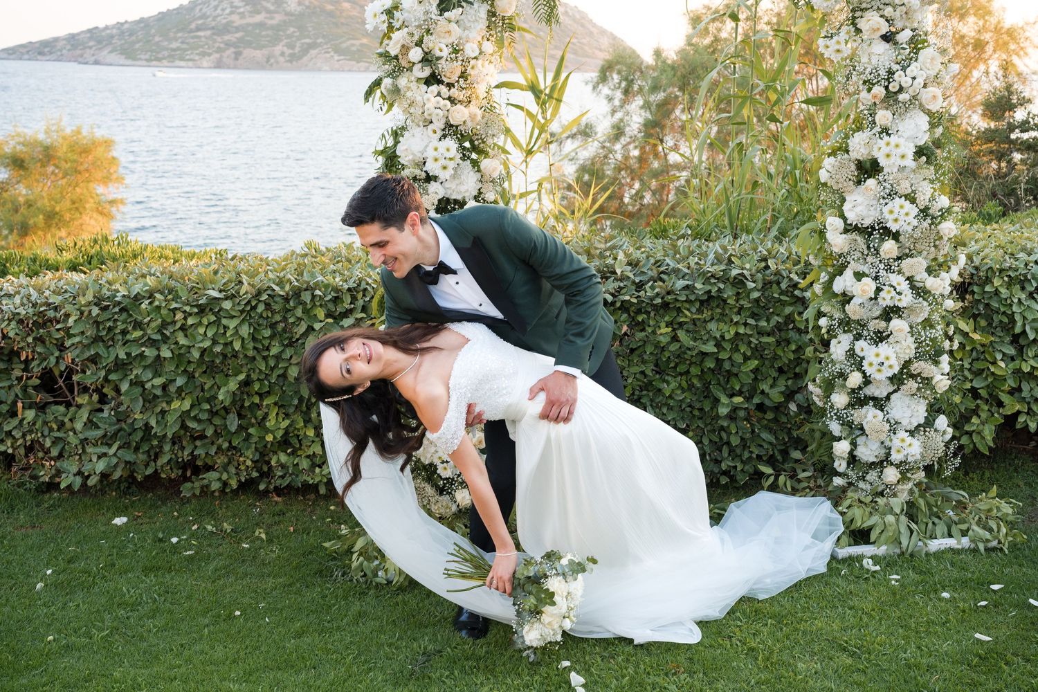 Bride and groom share a romantic dip kiss in front of a floral arch with ocean views at an outdoor wedding.
