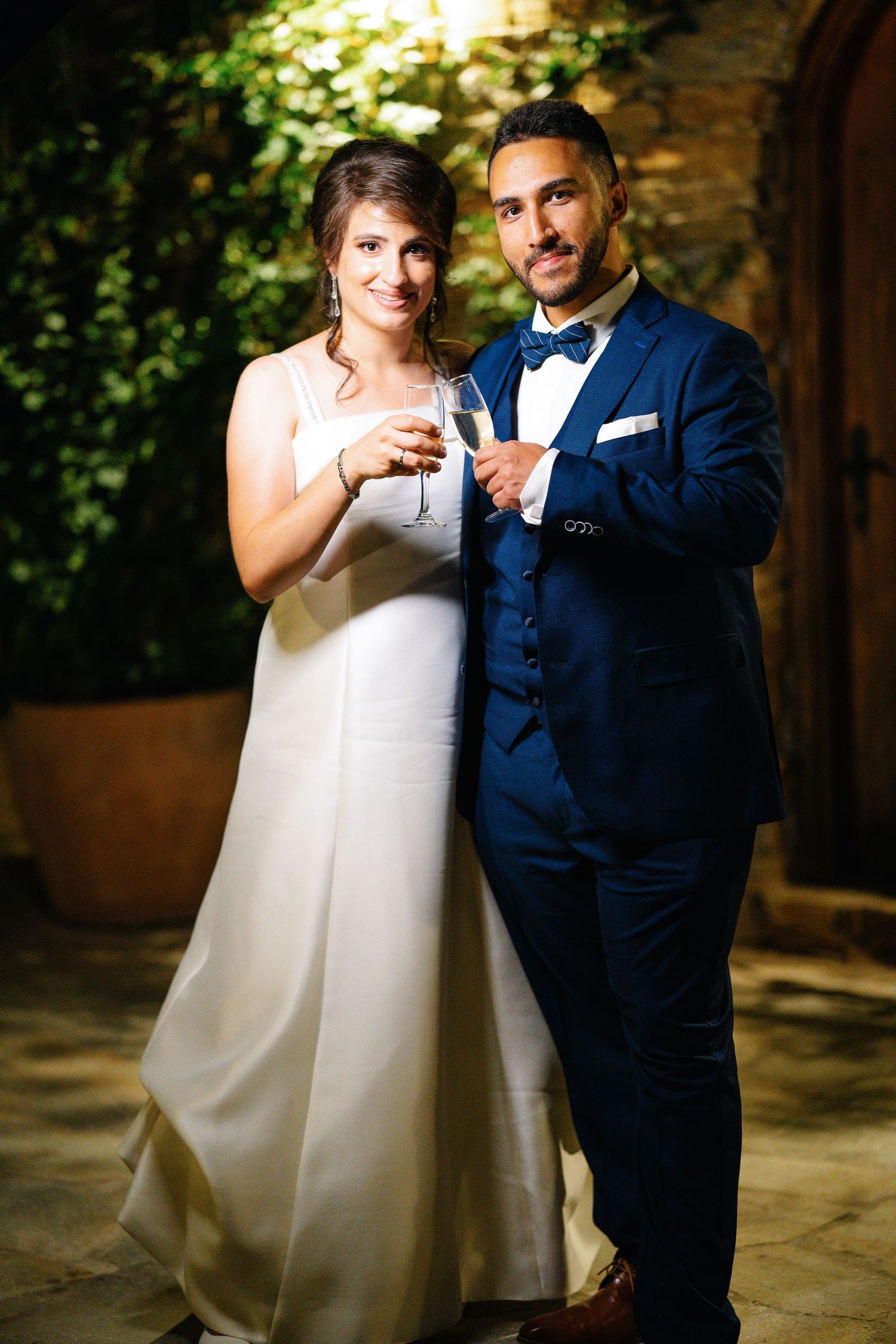A wedding couple poses intimately together in a garden setting with backlit foliage and rustic stone walls.
