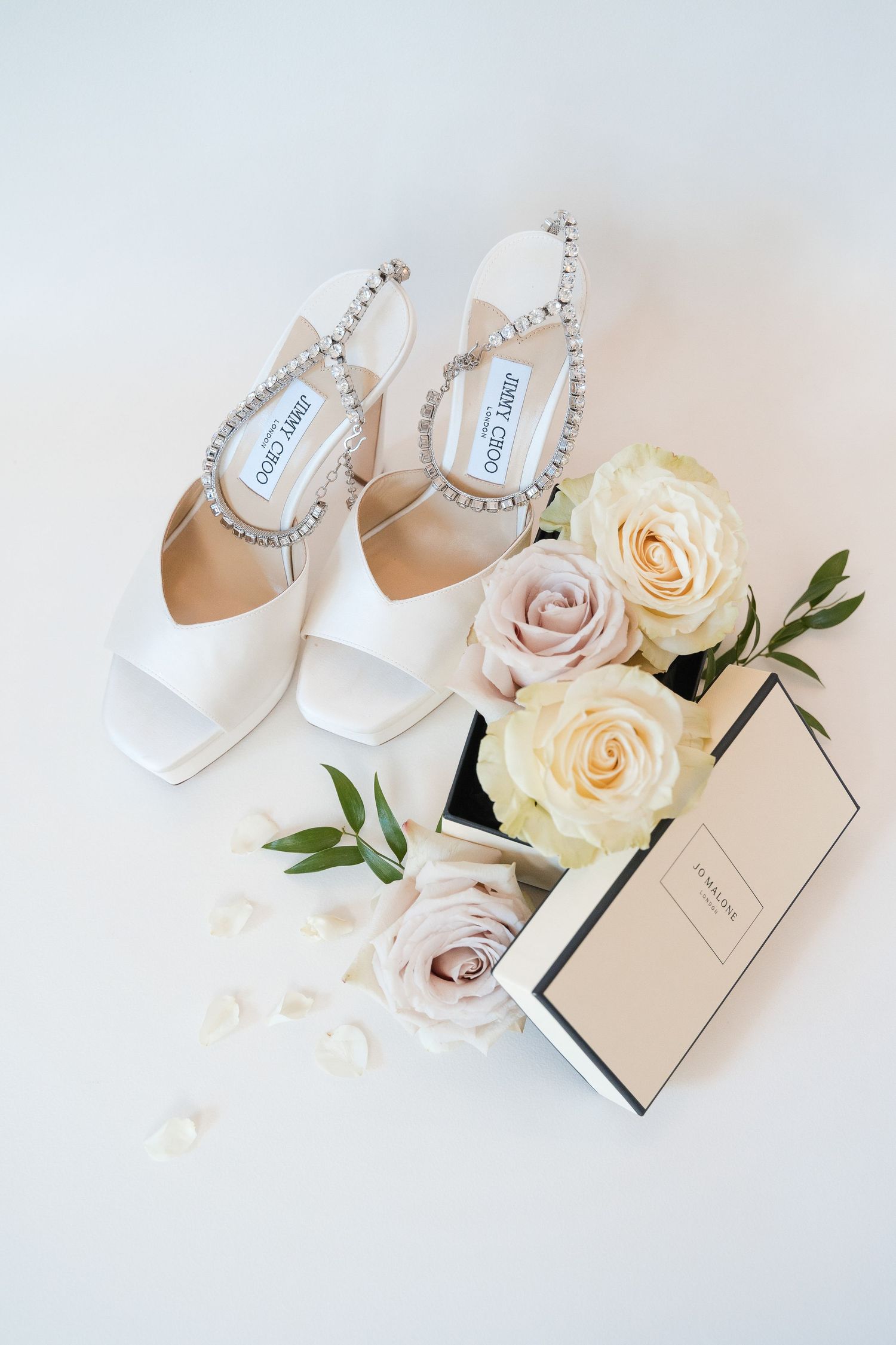 Elegant white Jimmy Choo bridal shoes with crystal straps displayed alongside blush and cream roses and a gift box.