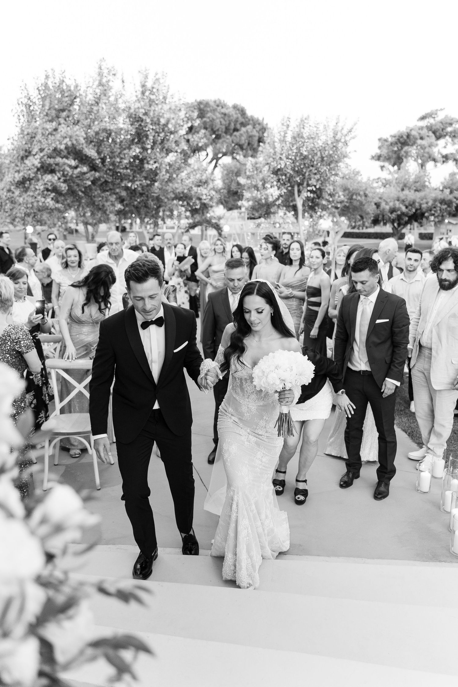 Black and white photo of wedding processional walking down outdoor aisle with guests.
