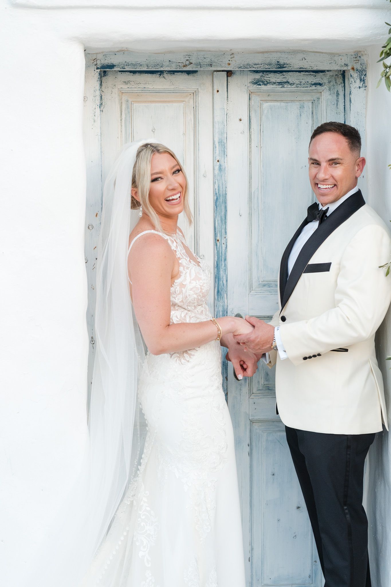 Joyful wedding portrait in front of weathered blue doors on a white Mediterranean building.