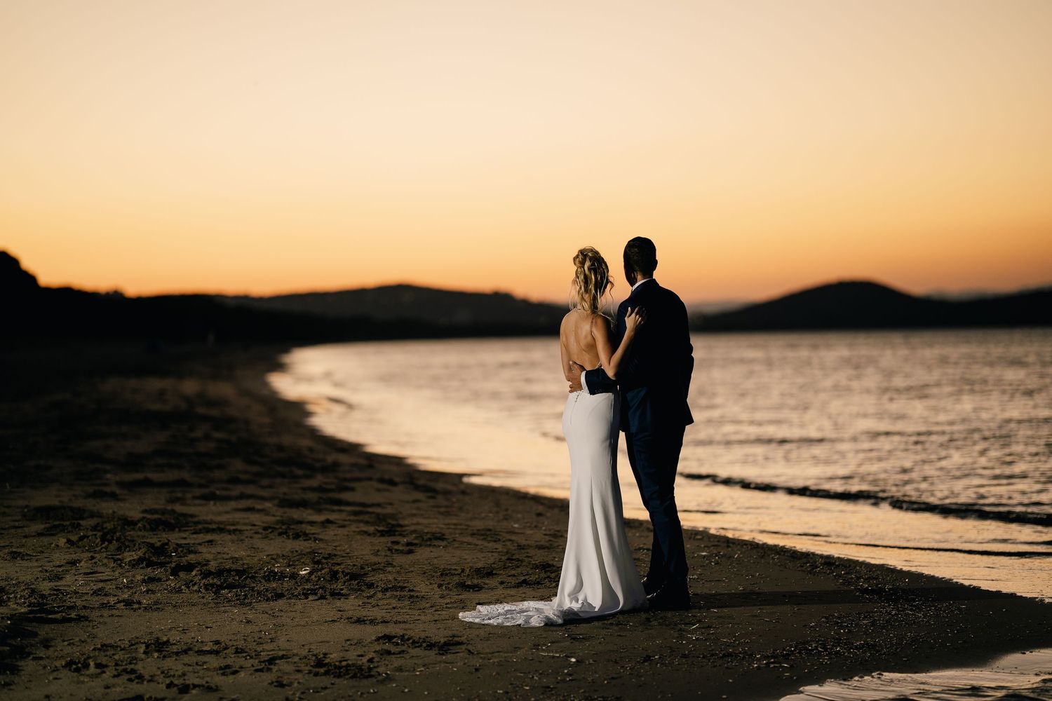 A couple in wedding attire stands silhouetted against a dramatic sunset on a beach shore.