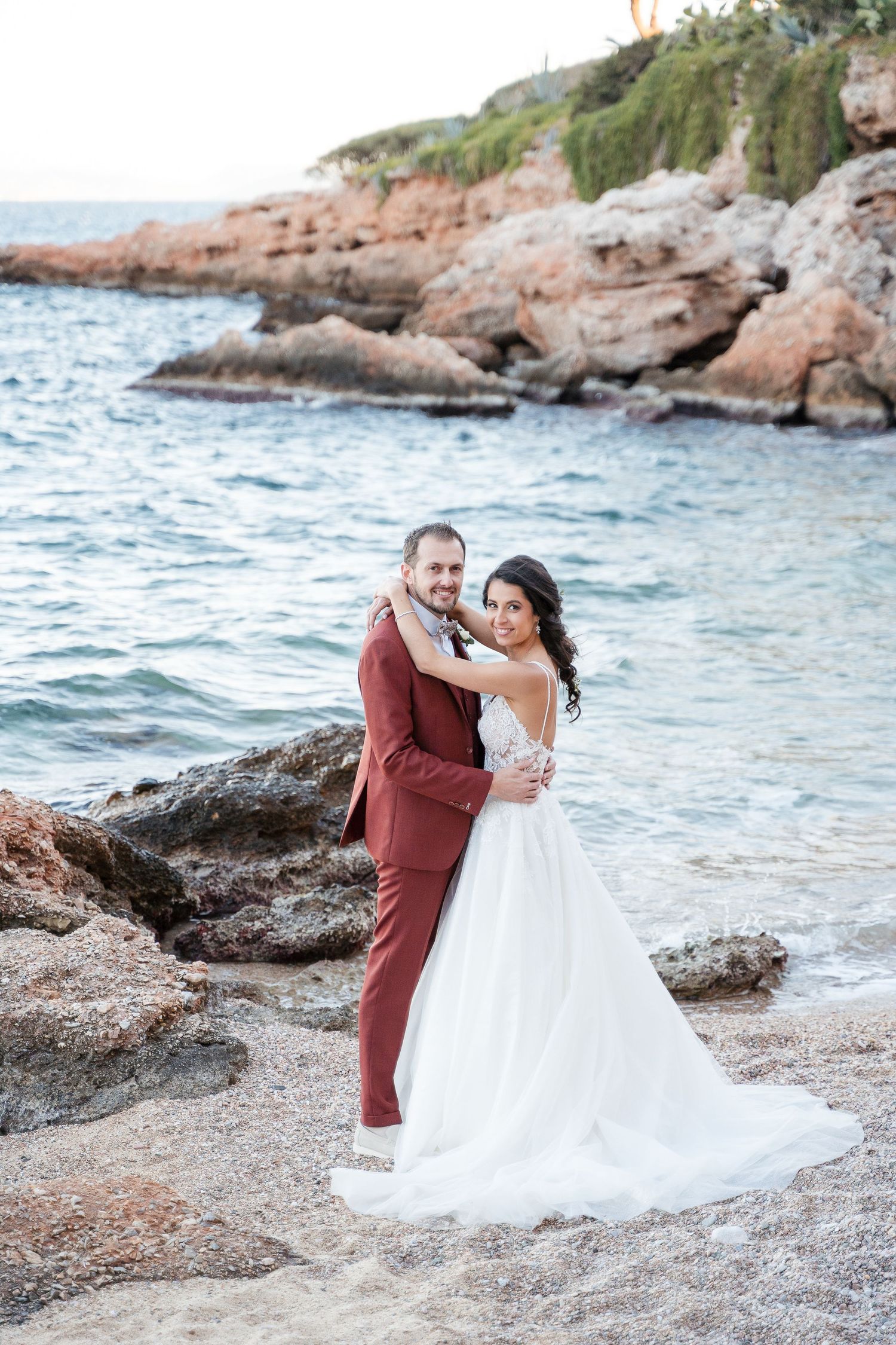 Newlyweds share intimate dance poses on rocky beach during sunset photography session.