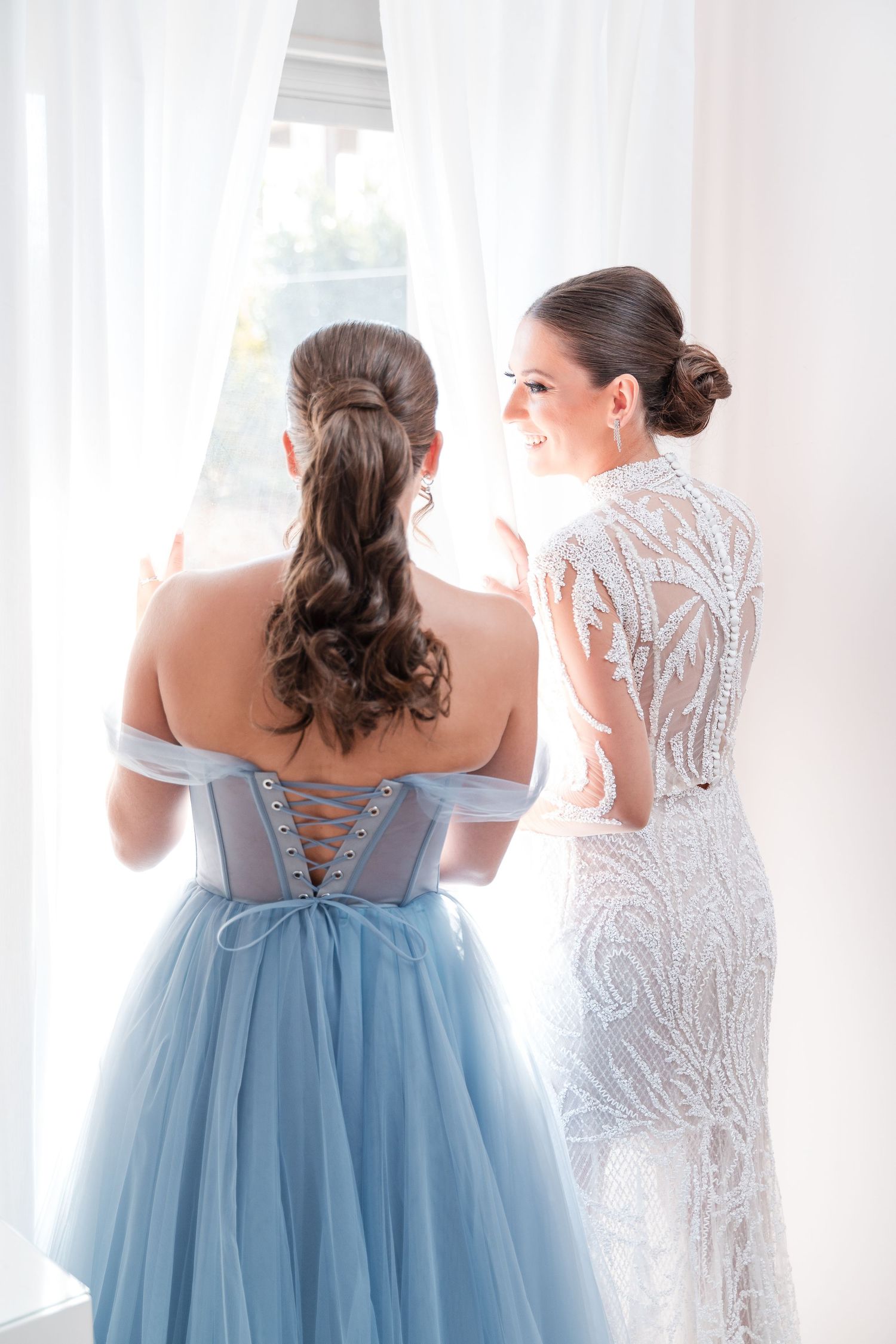 Two formal dresses shown from behind - one blue ballgown and one white beaded gown by window.