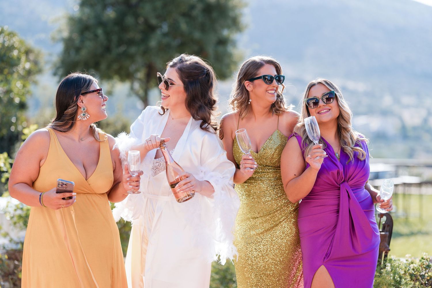 A group in colorful formal dresses including orange, white, gold and purple laugh together outdoors on a sunny day.