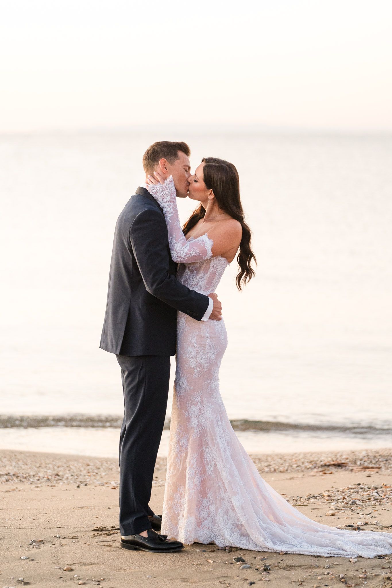 A couple shares romantic kisses on a sandy beach at sunset wearing formal wedding attire.