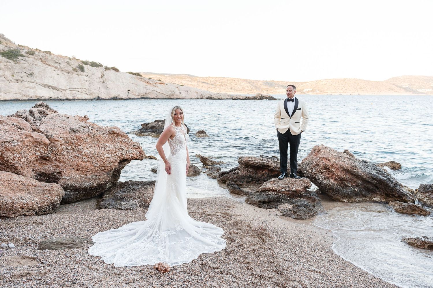 Bride in lace gown and groom in white tuxedo pose on rocky Mediterranean beach at sunset.