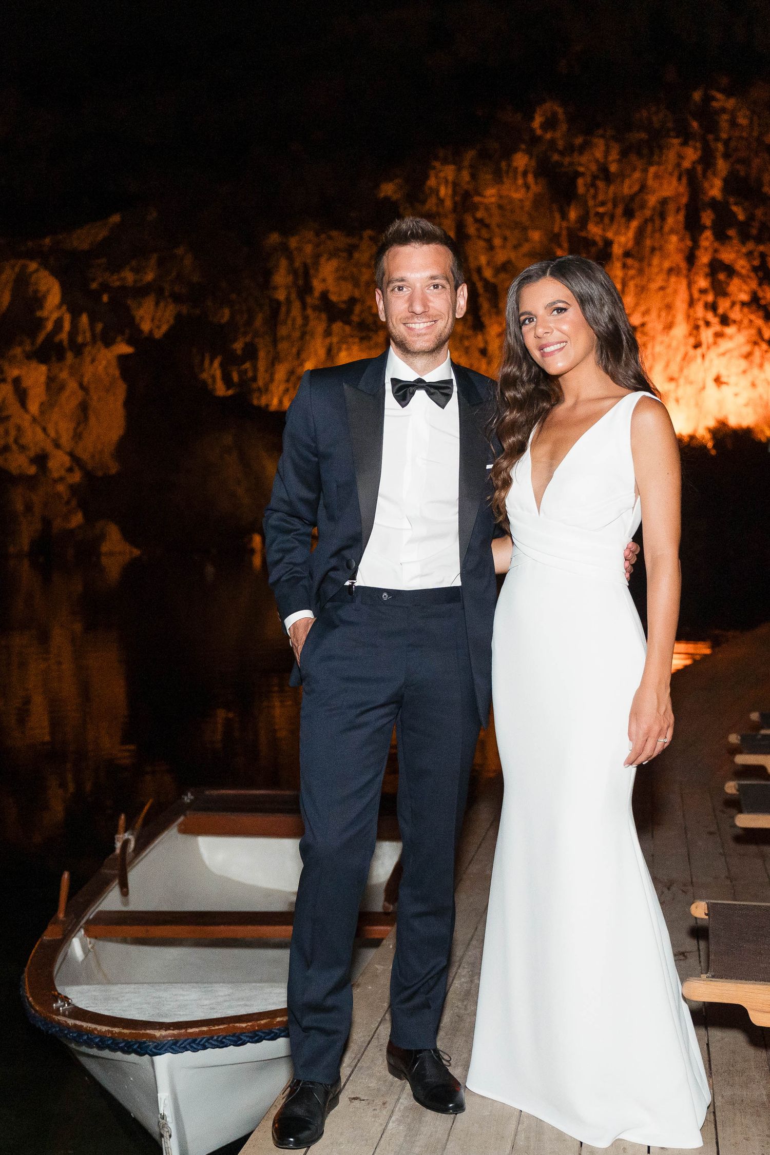 Formal wedding couple poses together at night in front of illuminated cave wall with boat in foreground.
