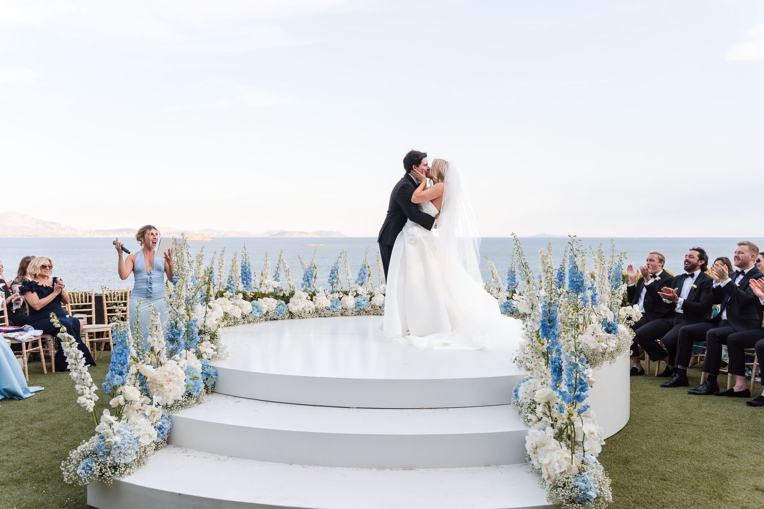 Bride and groom share first kiss on elegant circular white altar with blue floral arrangements at outdoor oceanview wedding.