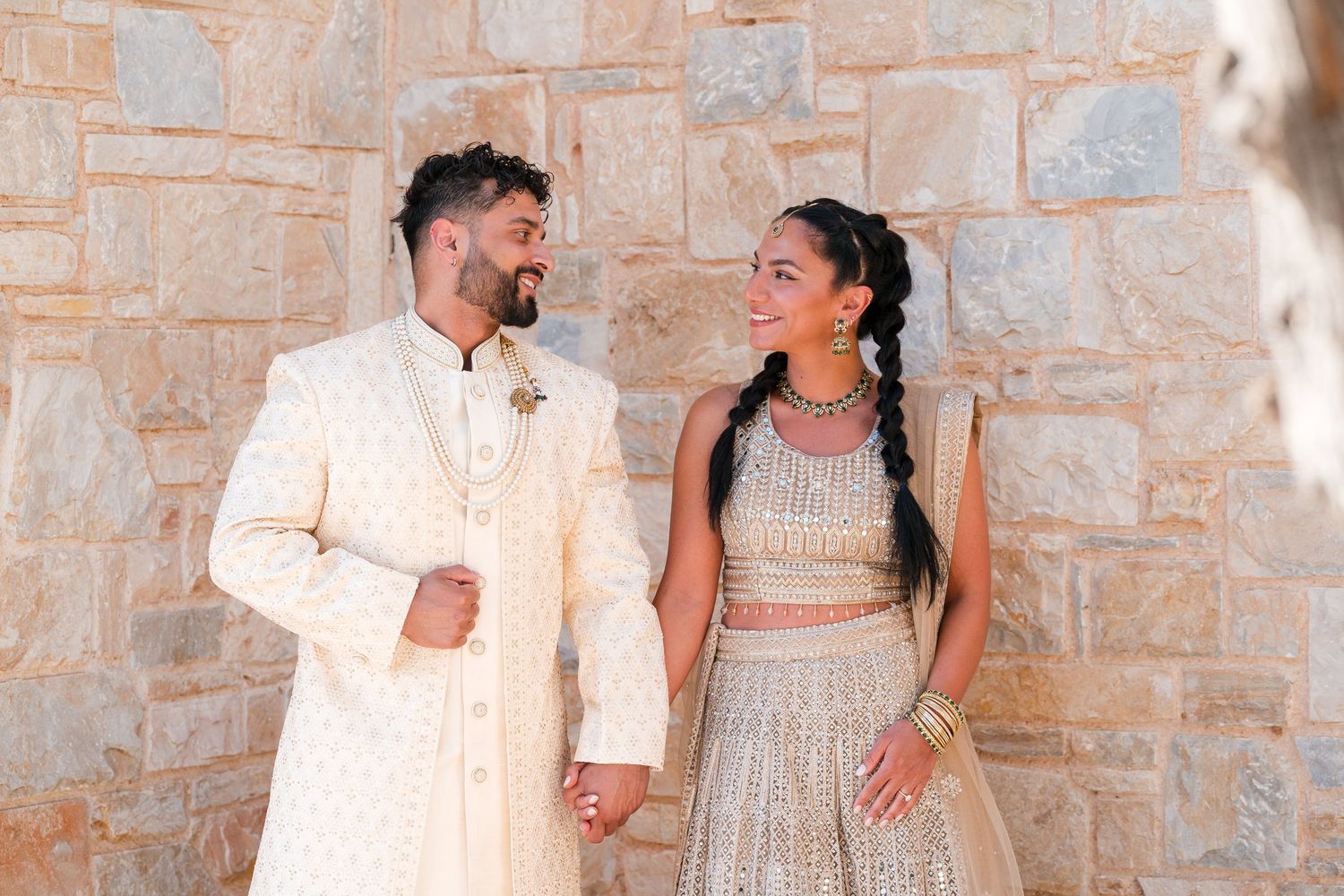 Indian couple in traditional wedding attire smiling at each other against a stone wall backdrop.