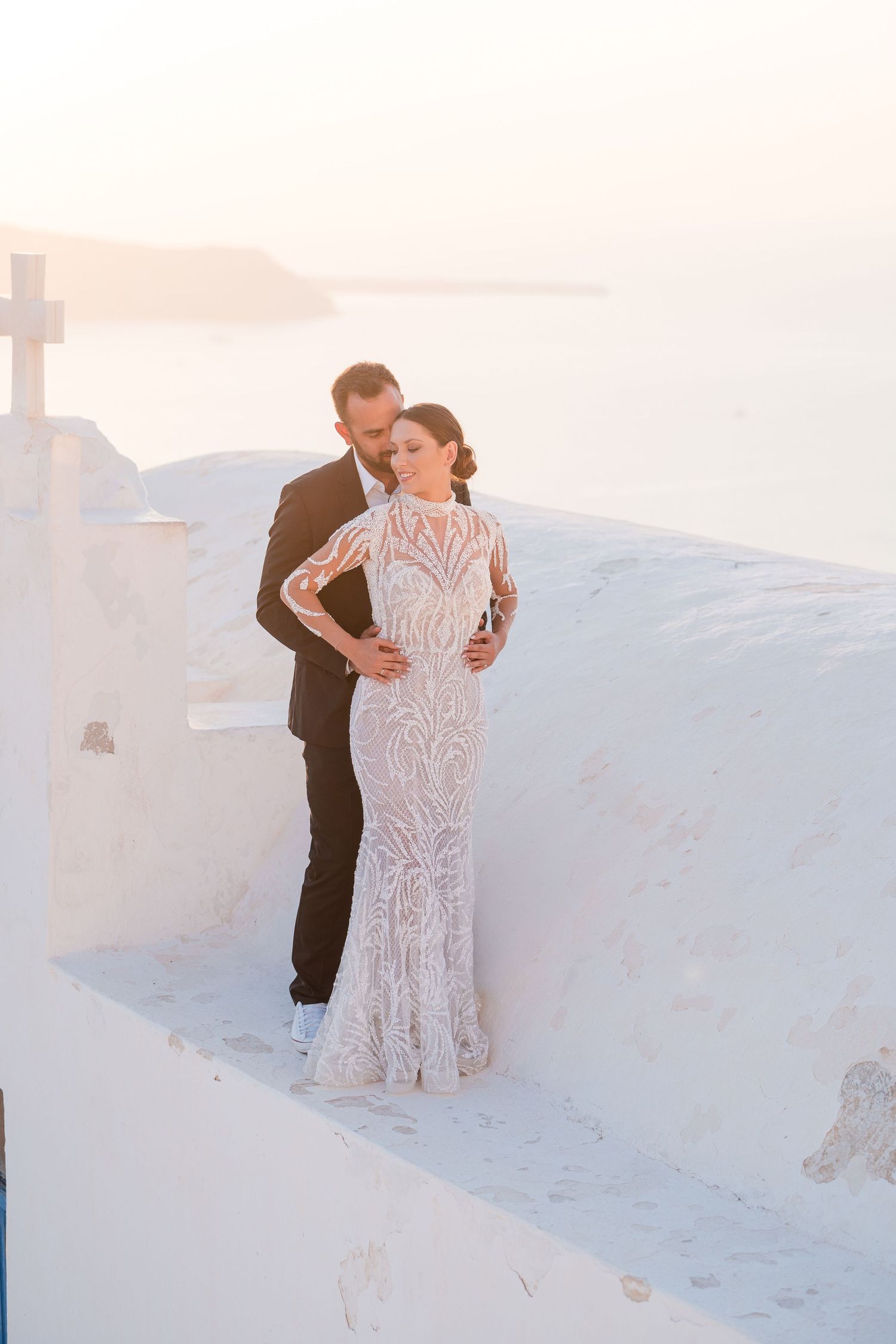 Couple embraces on sandy white hilltop during sunset in elegant lace wedding dress and black suit.