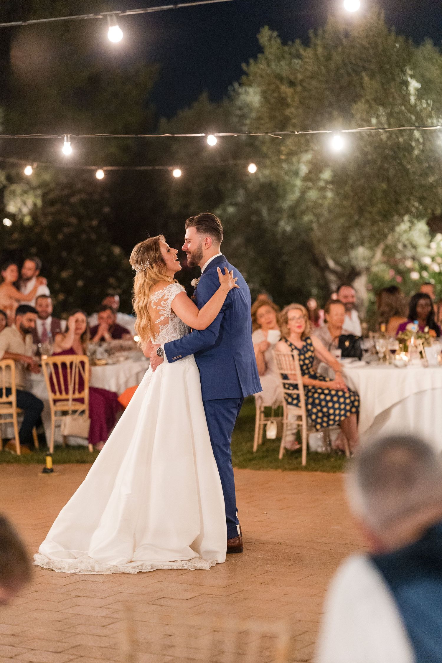 Romantic wedding couple shares first dance on outdoor patio under glowing string lights at evening reception.
