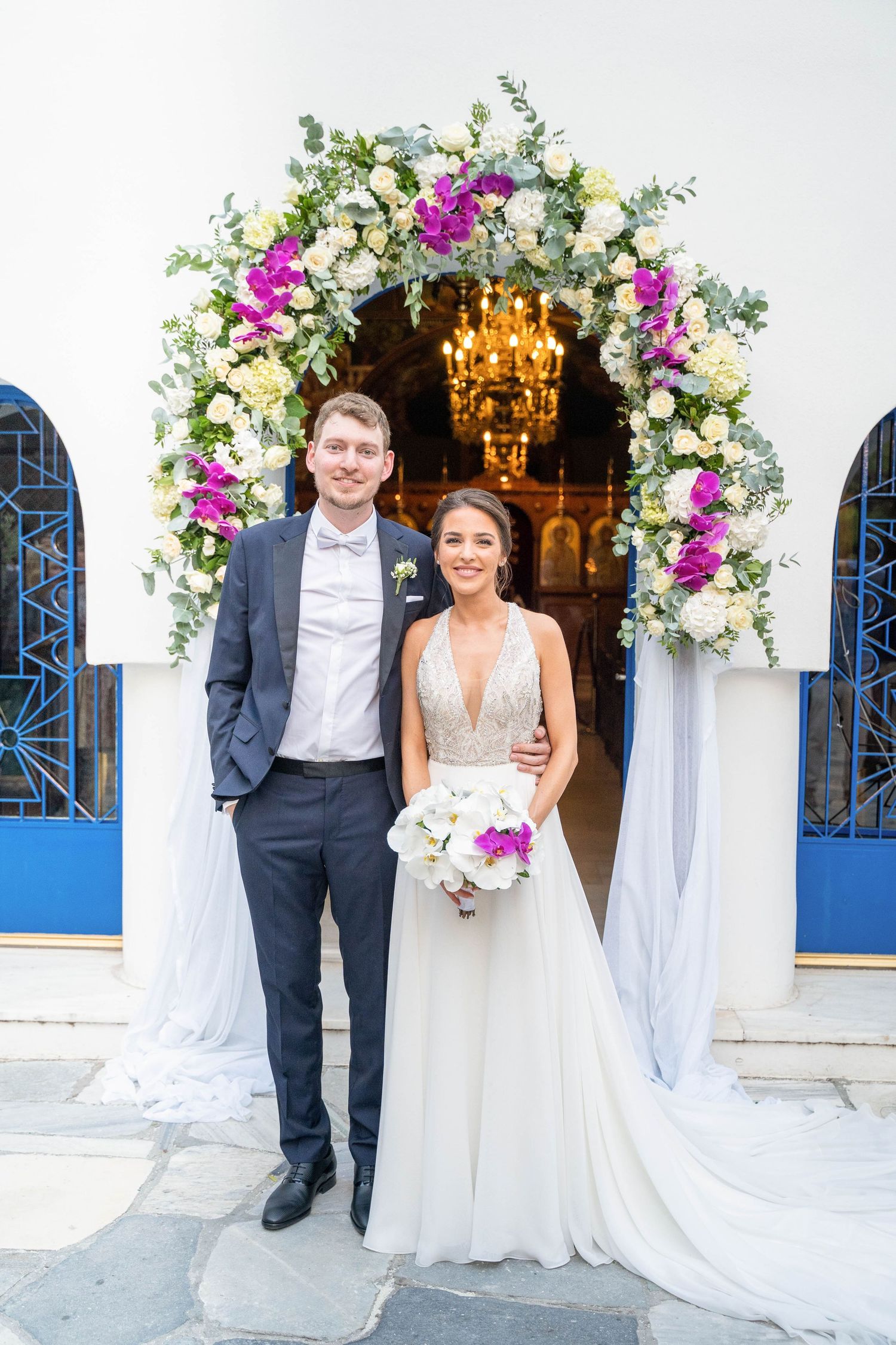 A wedding couple poses by a floral arch with pink and white flowers against a white wall with blue accents.
