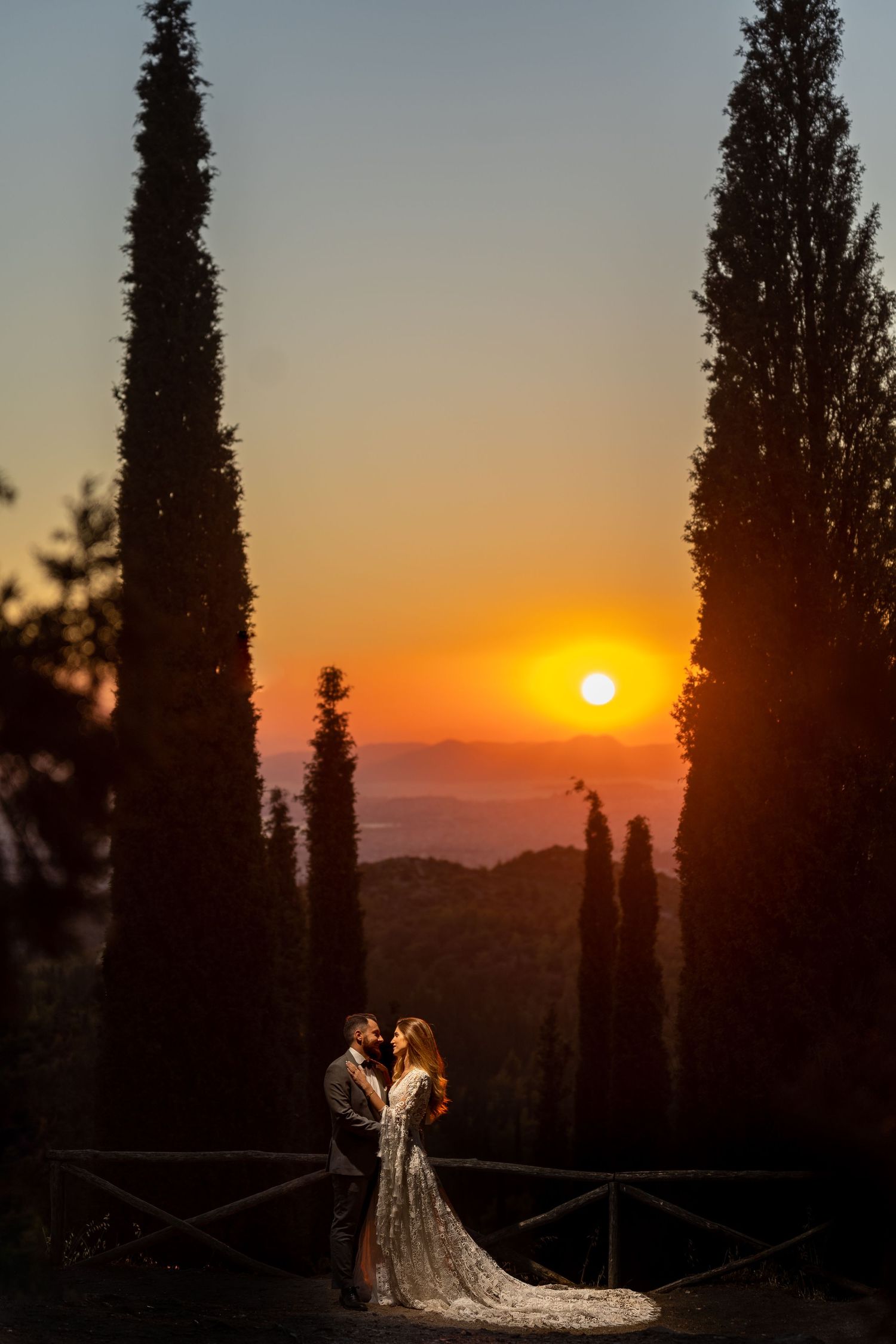 A spectacular sunset silhouettes tall cypress trees as a couple celebrates their wedding in a romantic outdoor setting.