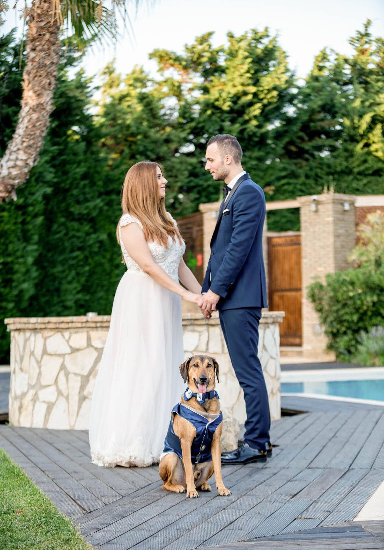 Sweet moment as couple embraces on wooden deck with their brown dog wearing blue bandana during outdoor wedding shoot.
