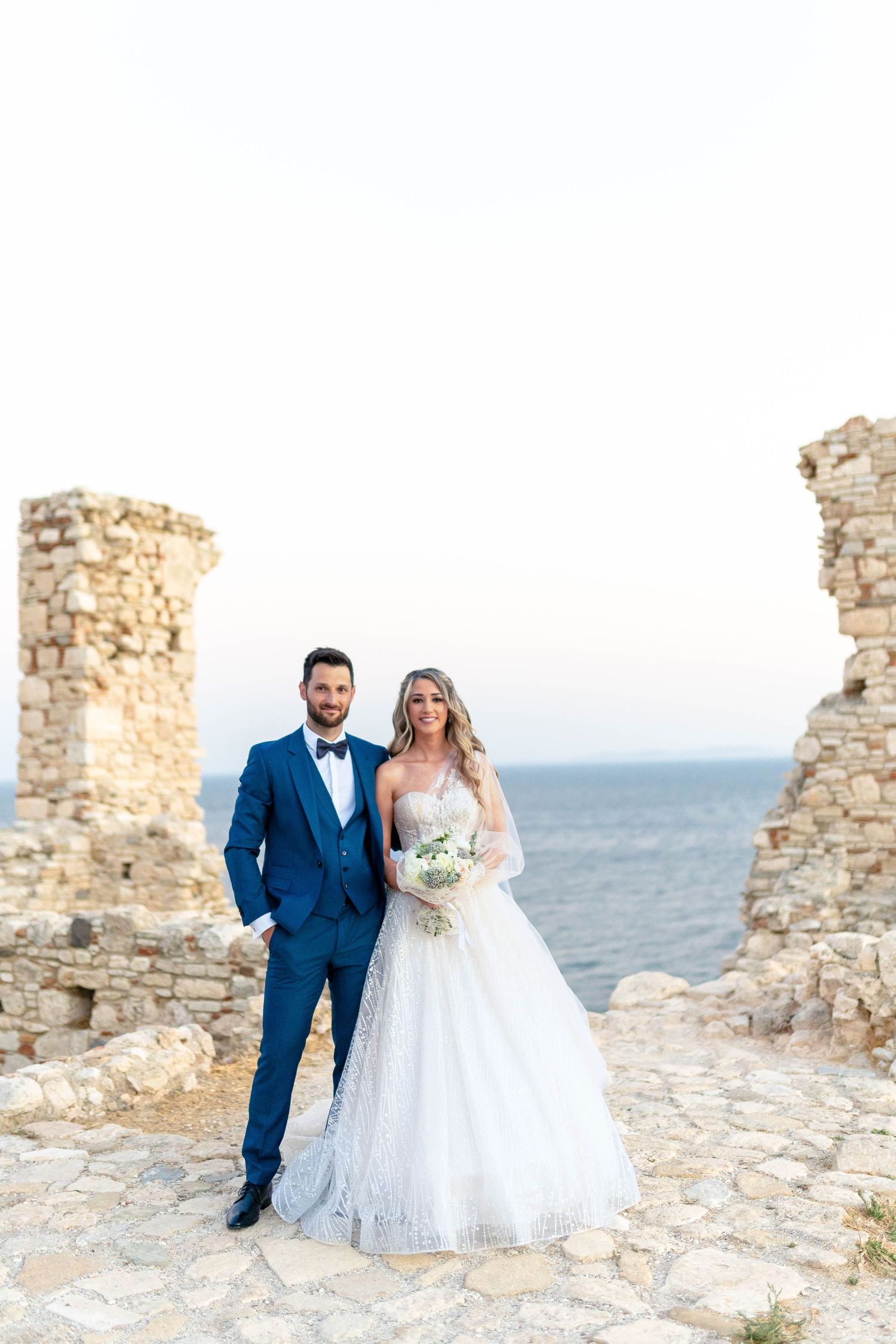 A couple poses at ancient stone ruins overlooking the ocean on their wedding day.
