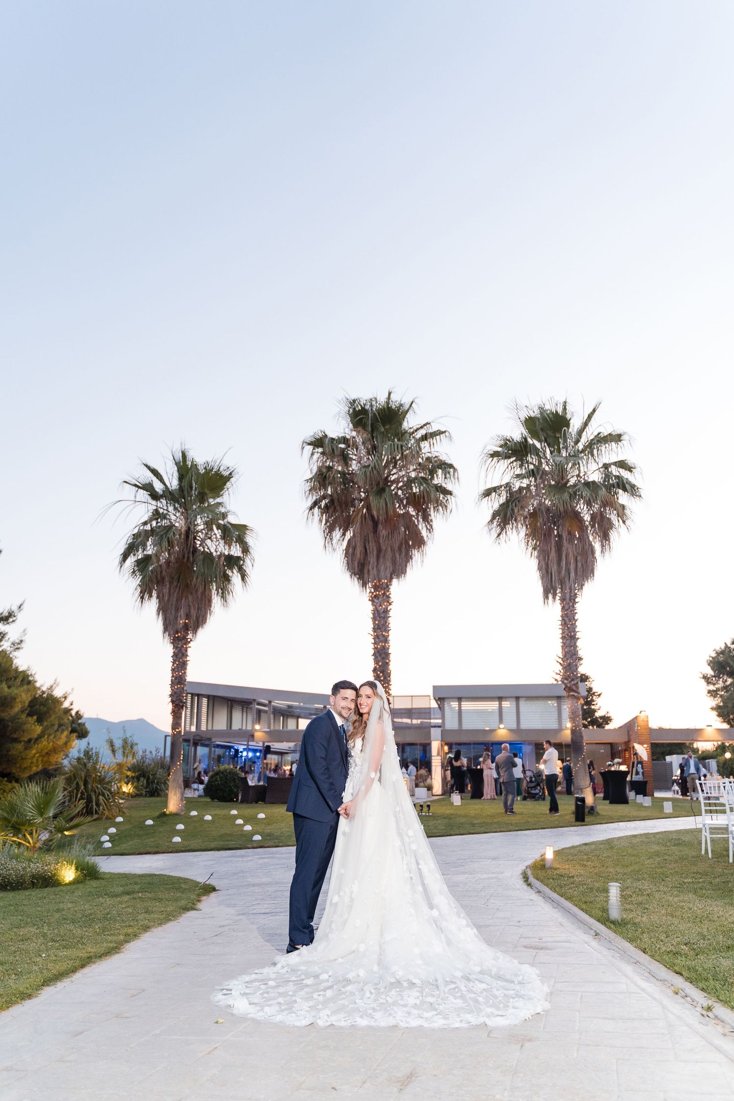 Bride and groom share an elegant moment on a grassy lawn with palm trees and waterfront views at sunset.