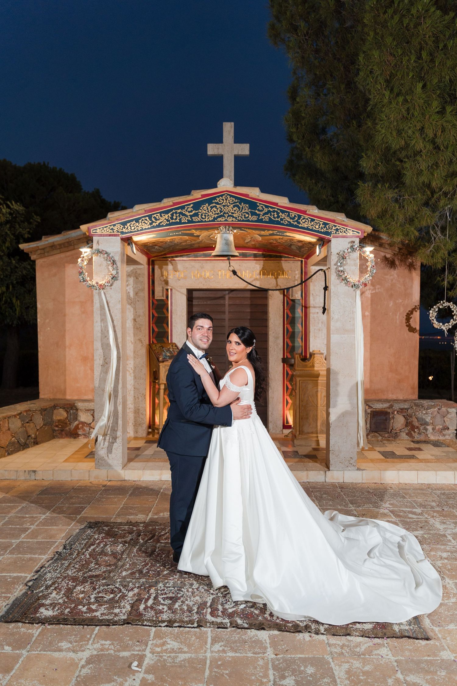 Romantic wedding couple poses in front of a stone chapel with adobe walls and white columns at dusk.