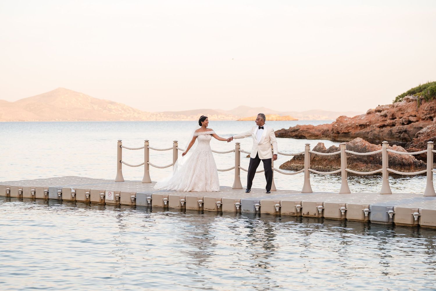 Bride and groom hold hands on a waterfront pier at sunset with rocky coastline and calm sea views.