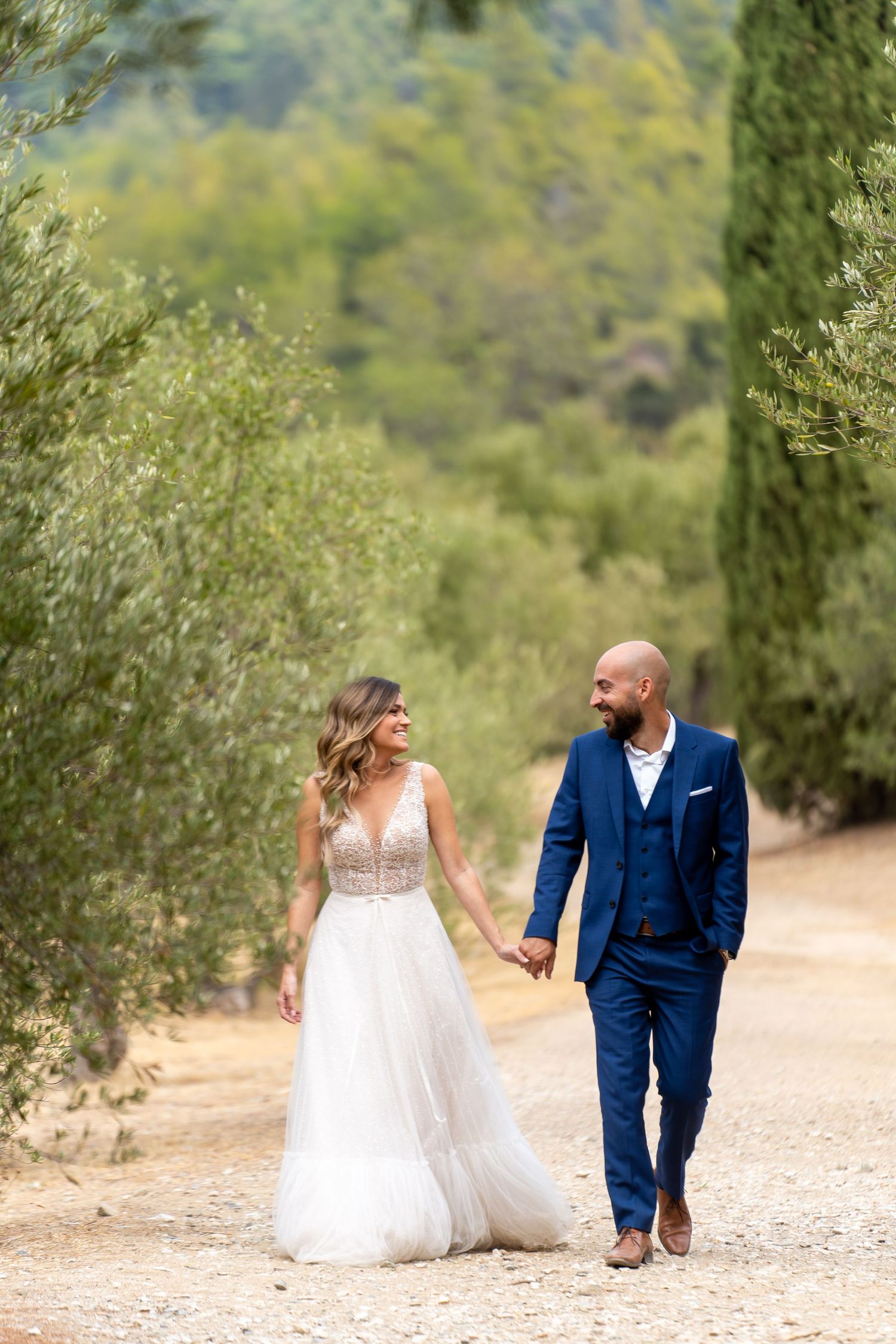 A couple in wedding attire walks along a scenic dirt path surrounded by green trees and vegetation.