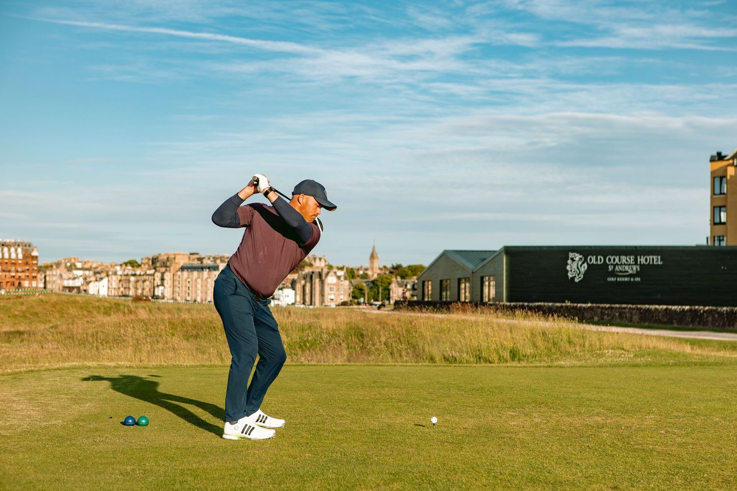 Golfer swings club on scenic coastal golf course, playing at Old Course.