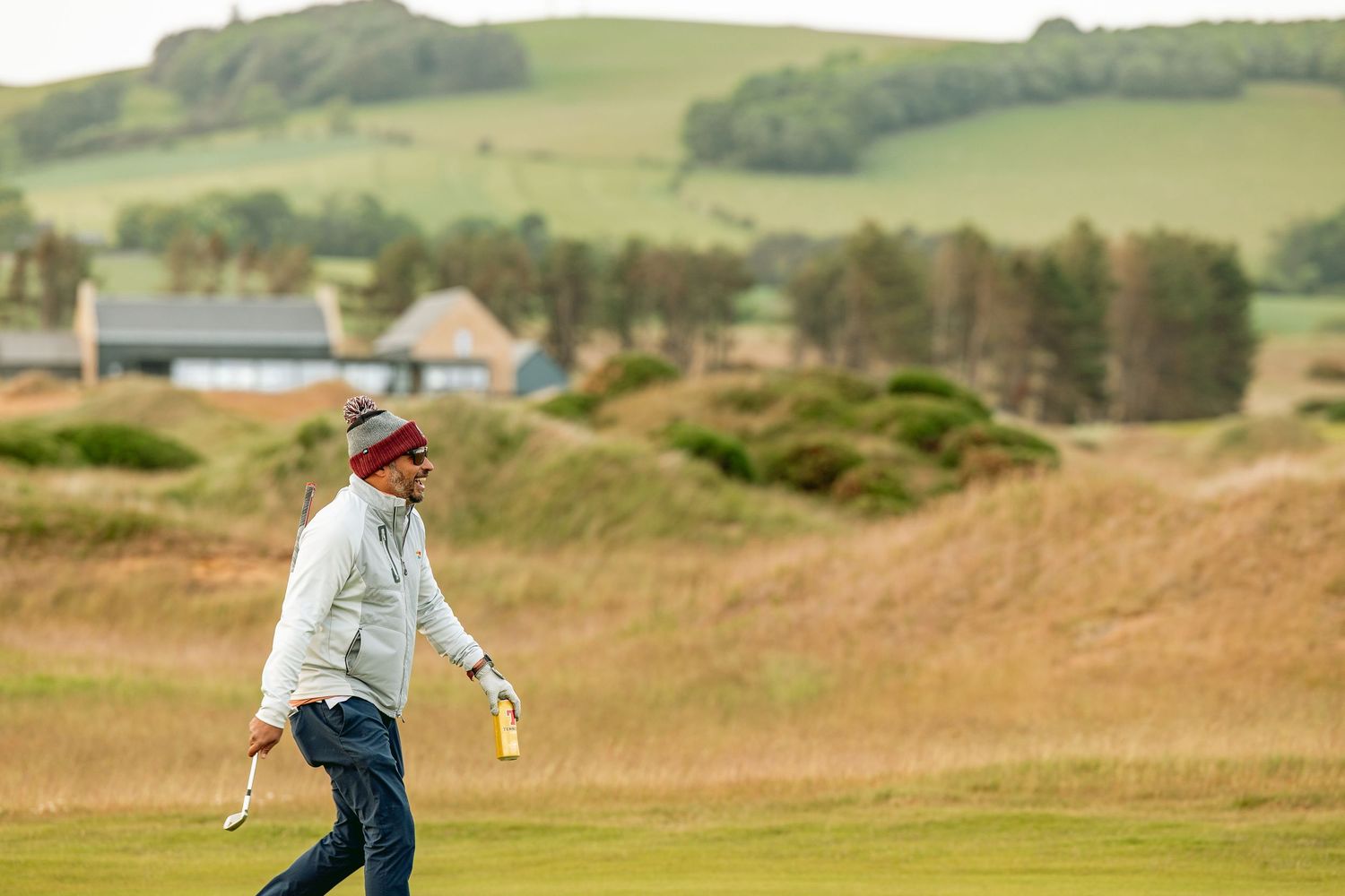 Happy golfer in white jacket, walking at golf course, Dumbarnie Links, with rolling terrain in background.