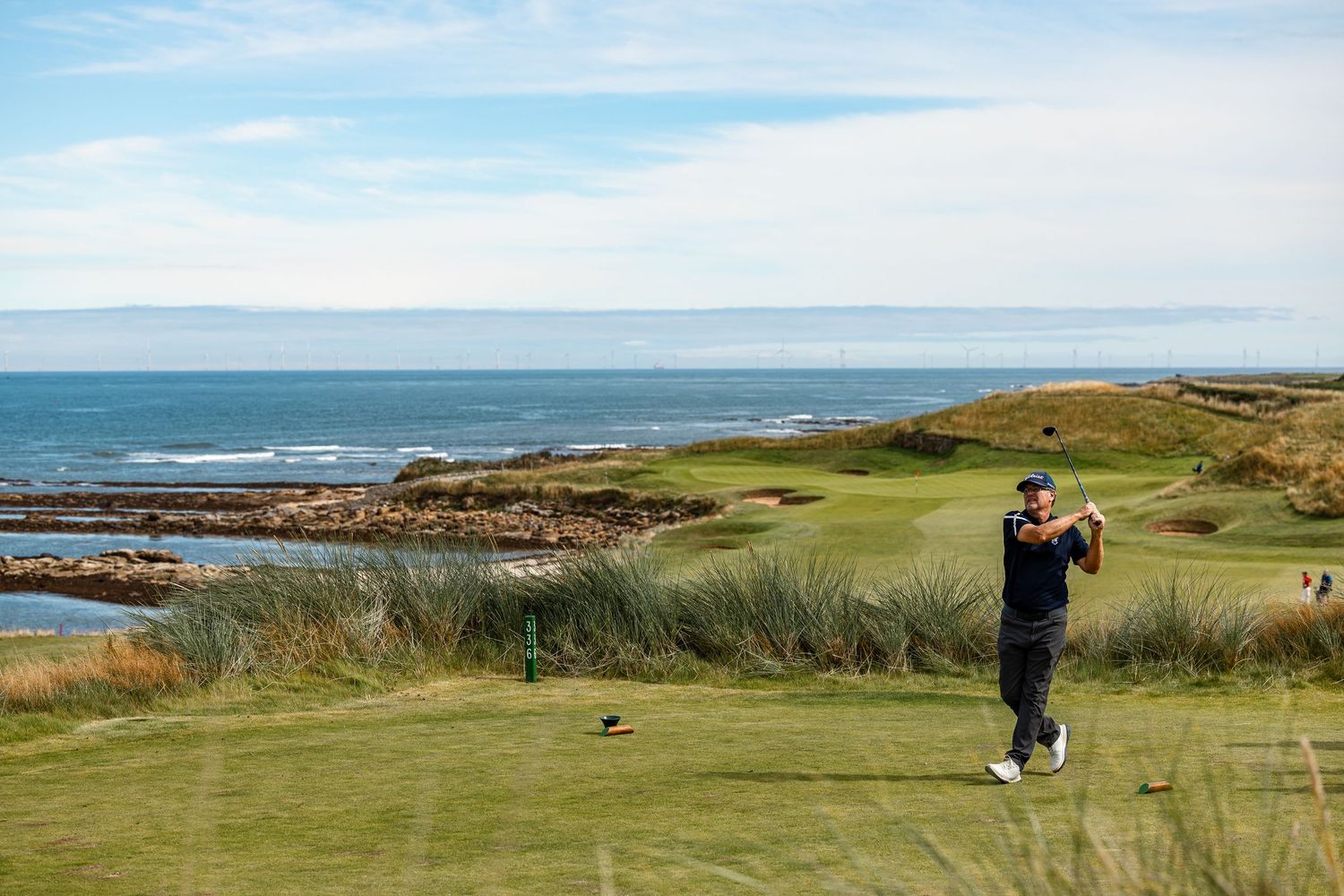 Golfer tees off on a stunning links course with rocky coastline and blue ocean stretching to the horizon.