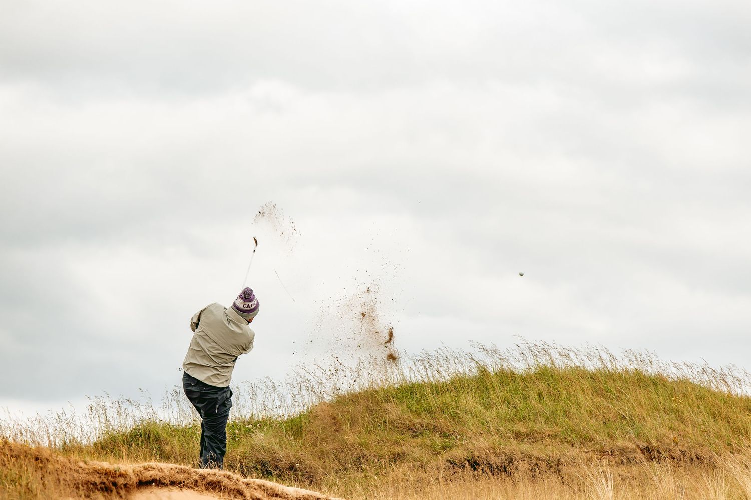A golfer swings golf club playing a big shot at Dumbarnie Links, against a cloudy sky.