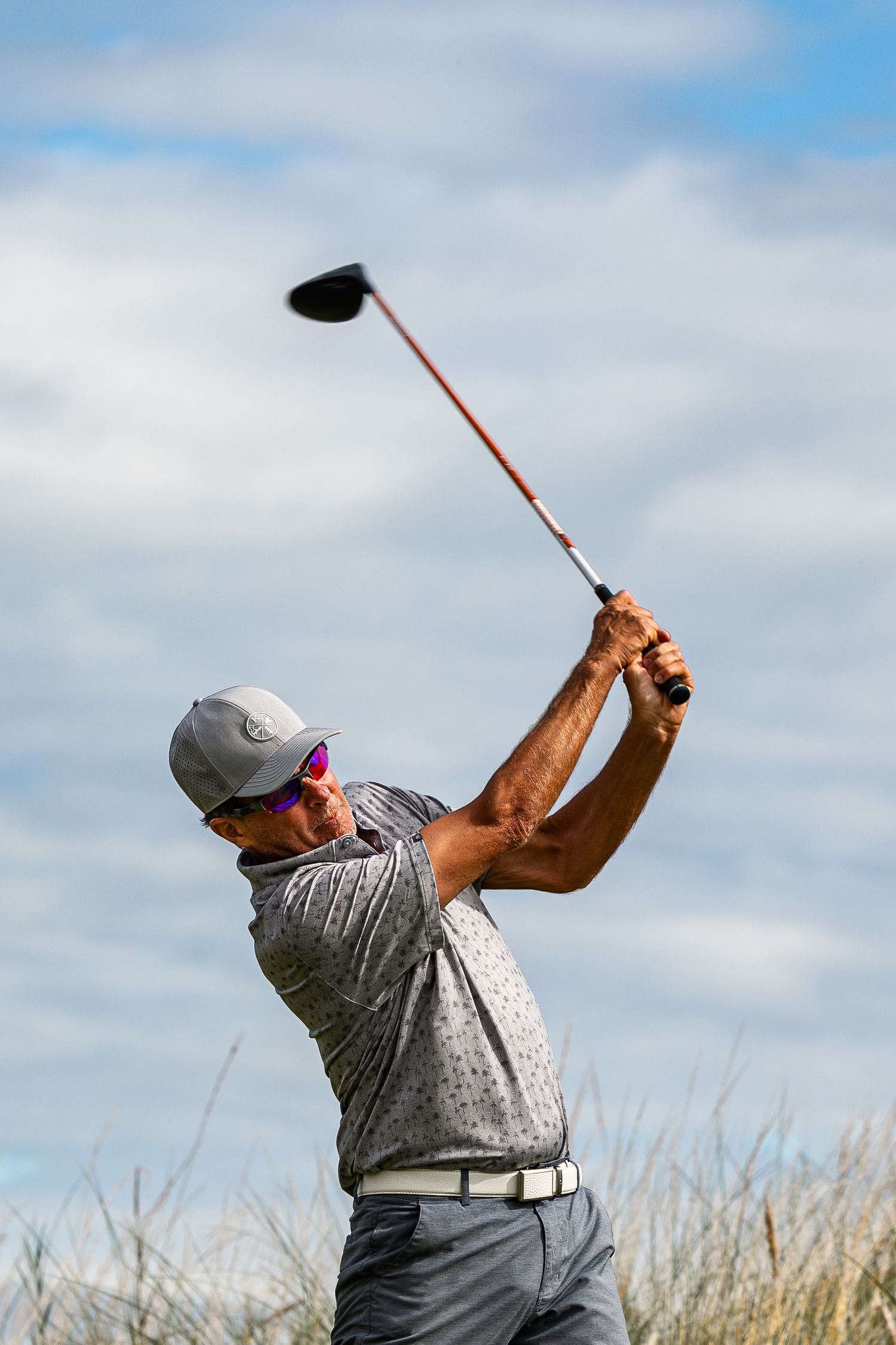 Golfer follows through on a powerful driver swing against a bright blue sky on a coastal links course.