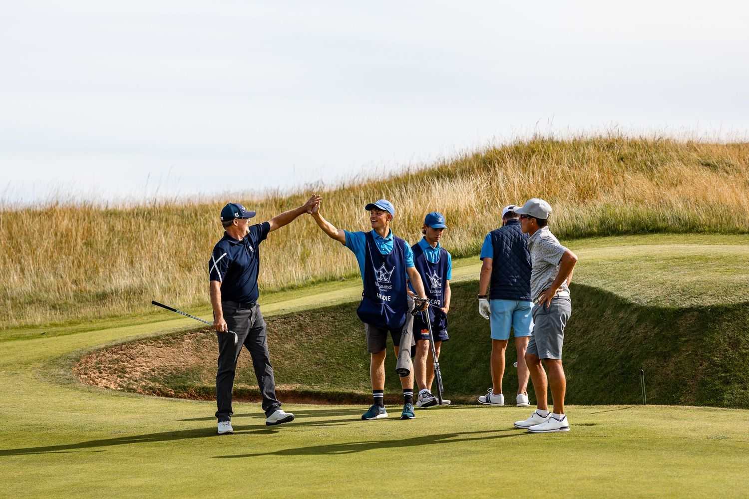 A golfer celebrates near a bunker as caddies and playing partners look on at a seaside links course.